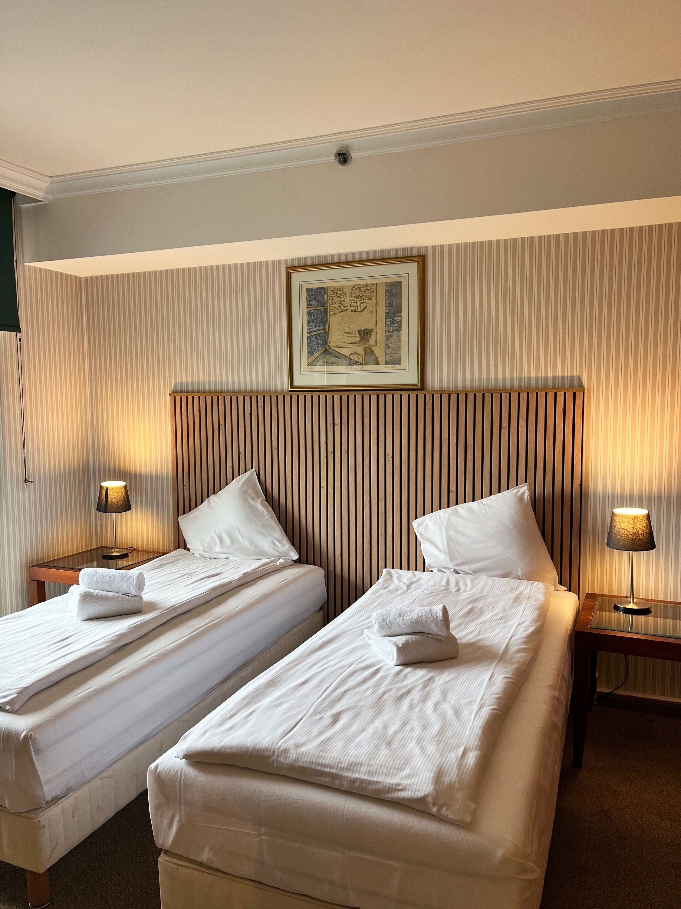 A hotel room with two single beds, each with a pillow and a rolled towel, a wooden headboard, a framed picture on the wall, and two bedside lamps.