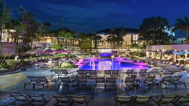 A resort pool area with lounge chairs and umbrellas, surrounded by palm trees and modern buildings. - Hilton Trinidad & Conference Centre (Port of Spain)