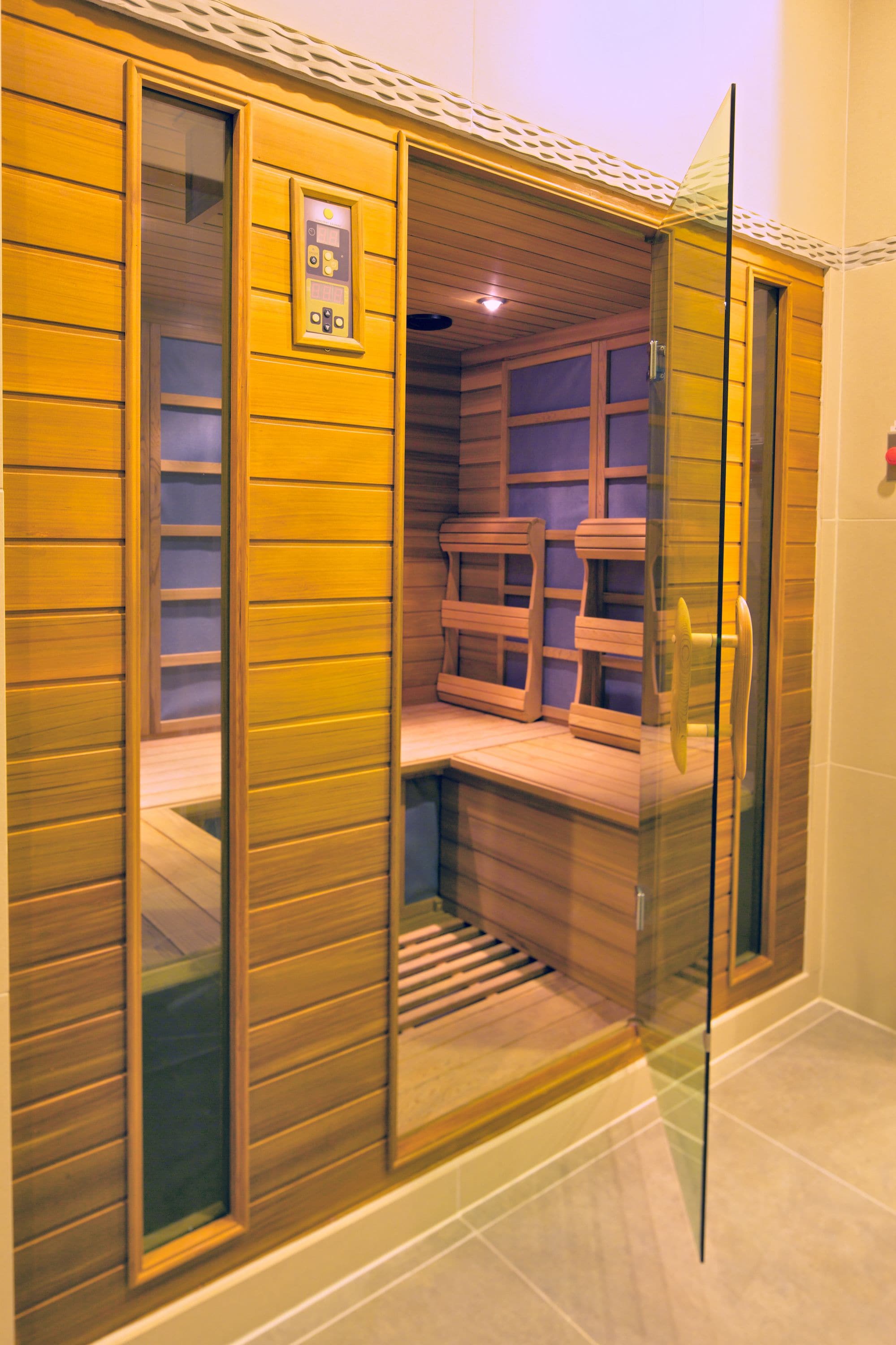 A sauna with wooden benches and a glass door, featuring a control panel on the wall.