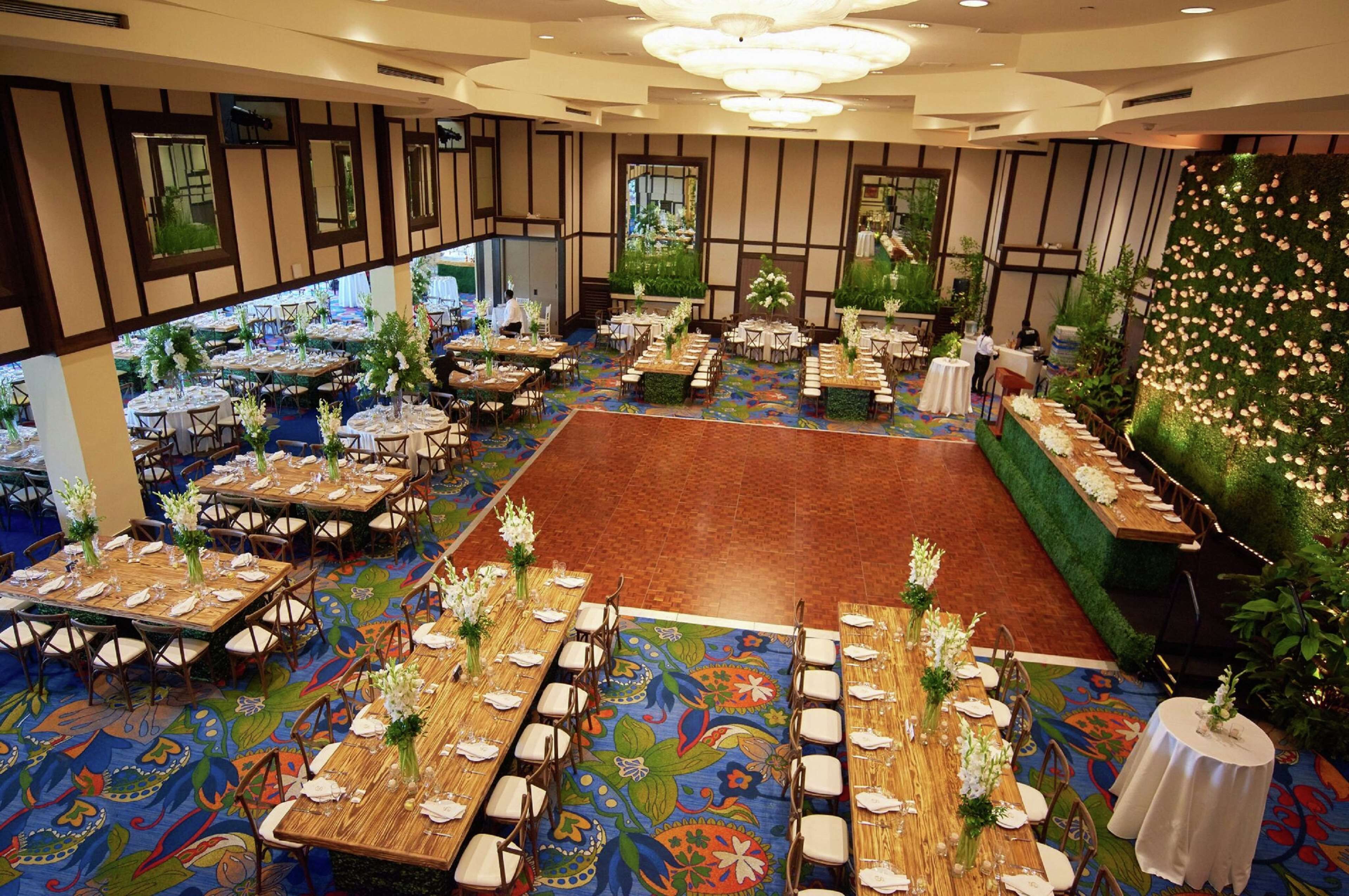 A banquet hall with multiple round tables, each set with white tablecloths and floral centerpieces. The room features a colorful carpet and a large, decorative wall with a floral arrangement.