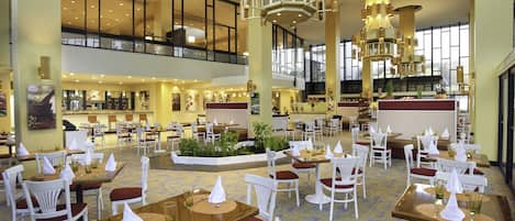 A spacious hotel lobby with multiple dining areas, high ceilings, and large windows.