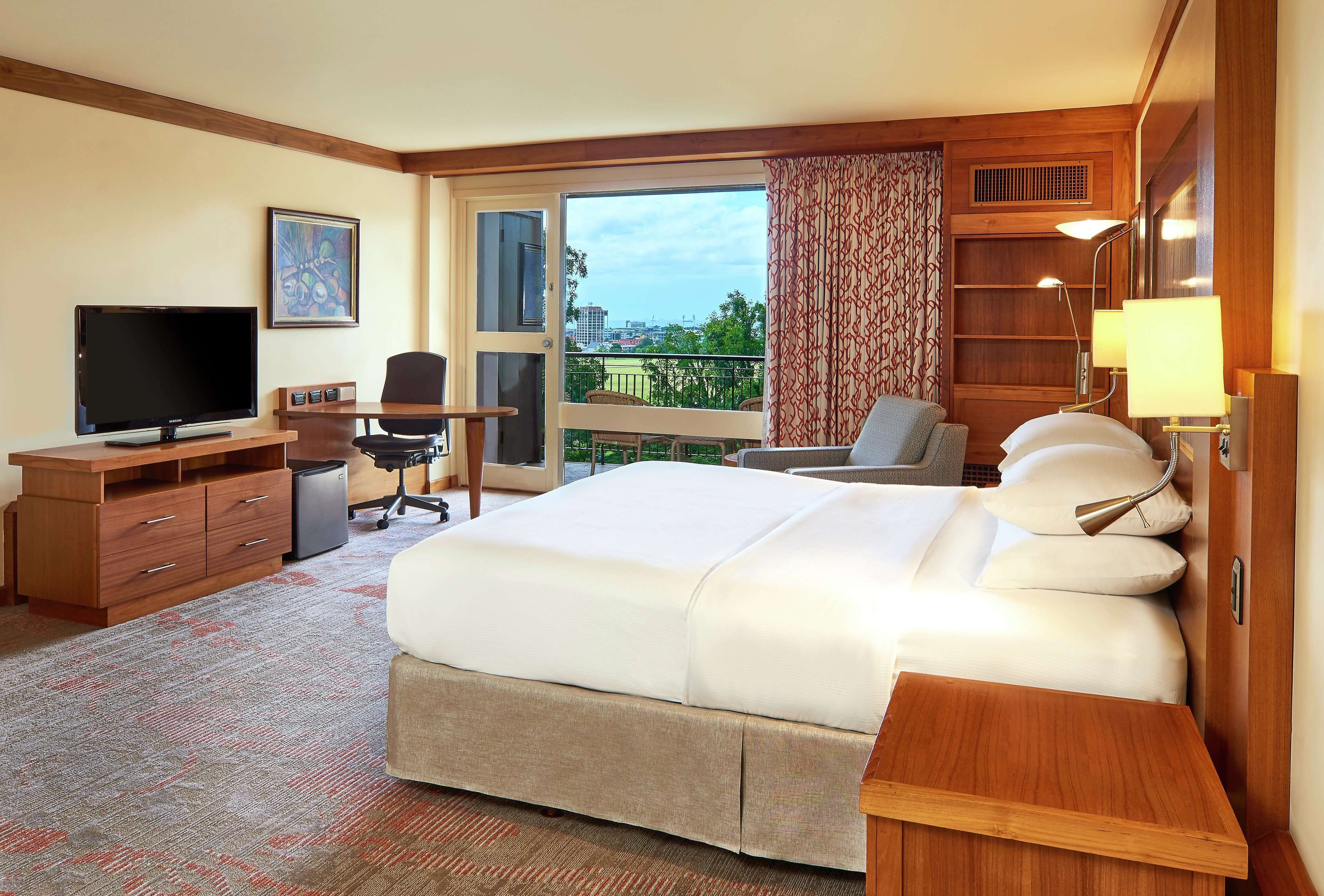 Habitación de hotel con una cama grande, un escritorio con televisión, una silla y vistas a la ciudad a través de una ventana.