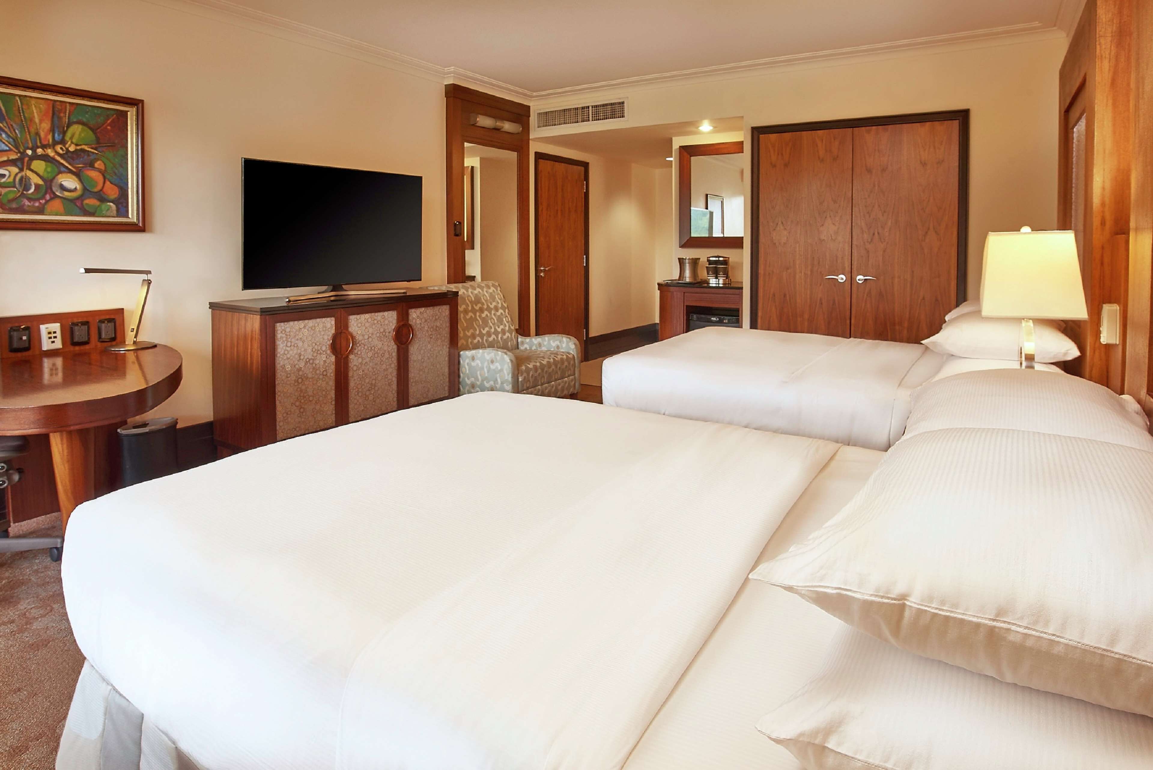 A hotel room with two beds, a television, a desk with a lamp, and a painting on the wall.