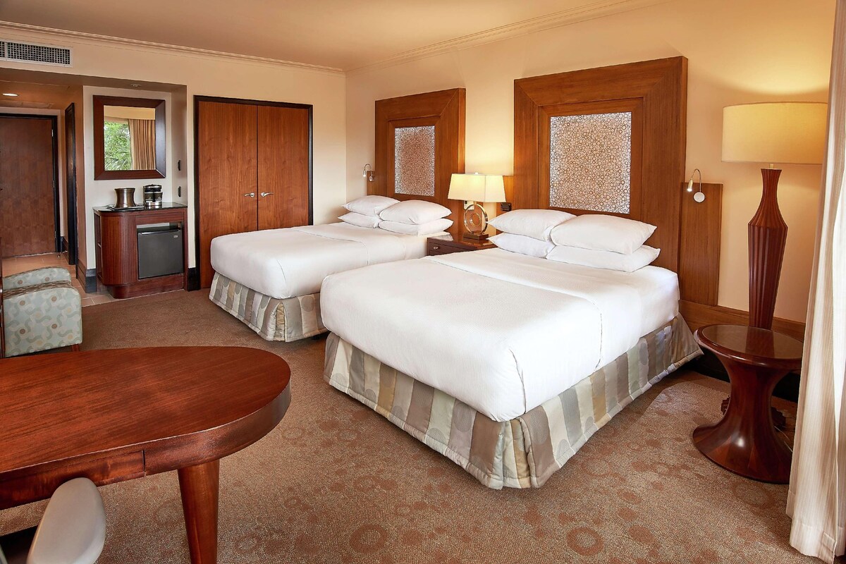 A hotel room with two beds, a desk, a chair, a small refrigerator, and a lamp.