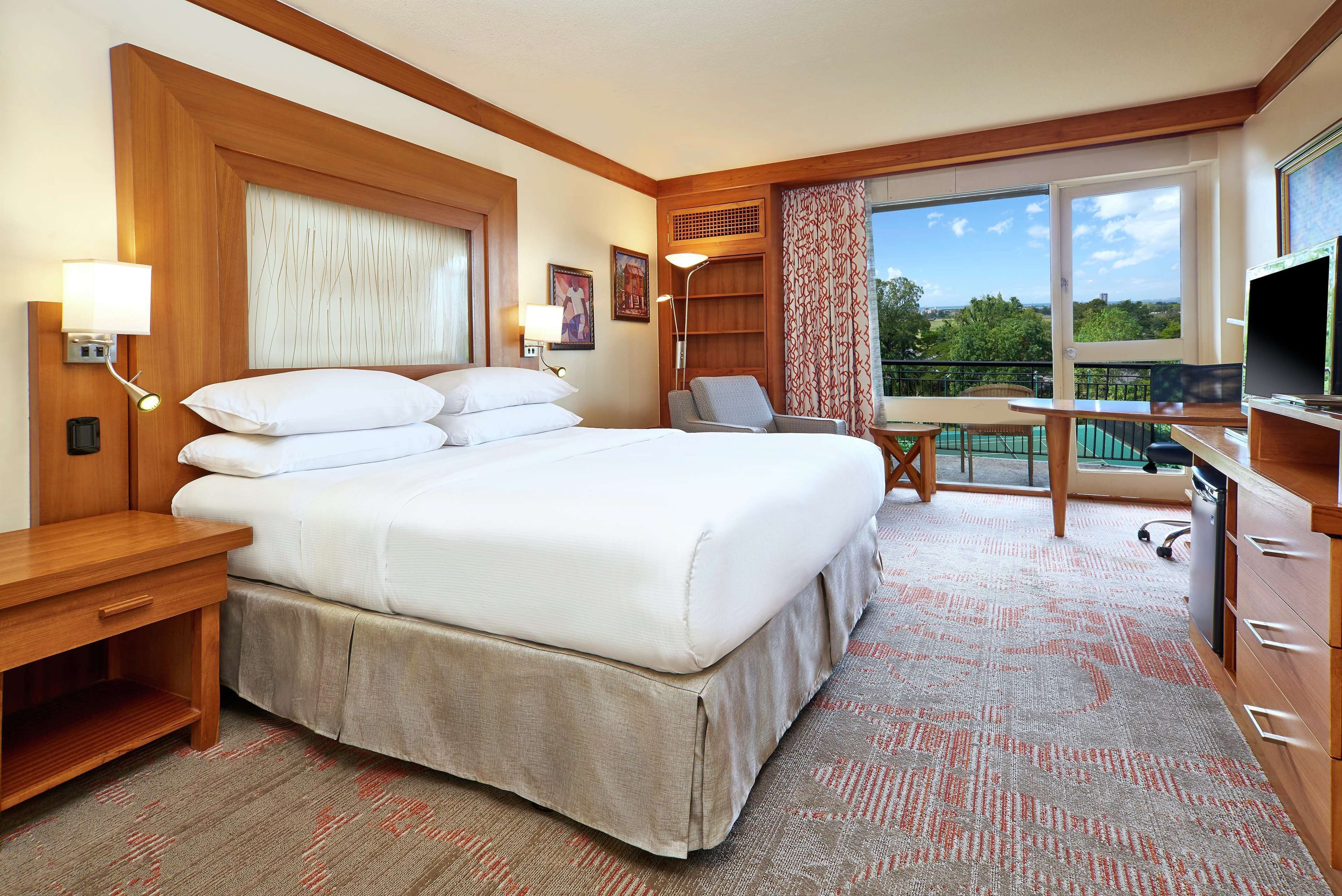 A hotel room with a large bed, a desk, a chair, a TV, and a balcony with a view.