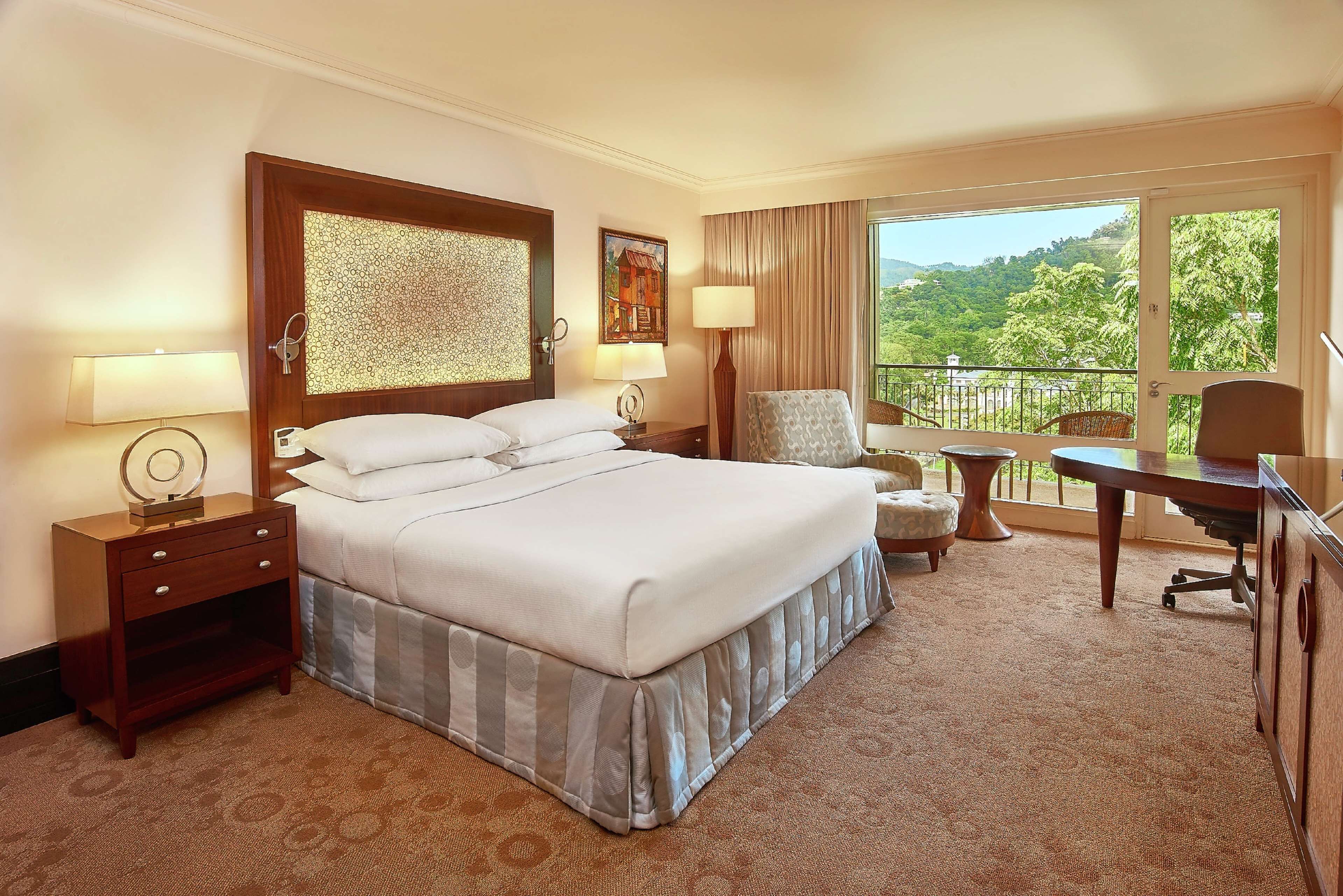 A hotel room with a large bed, a desk, a chair, and a balcony with a view of greenery.