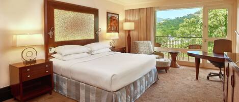 A hotel room with a large bed, a desk, a chair, and a balcony with a view of greenery.