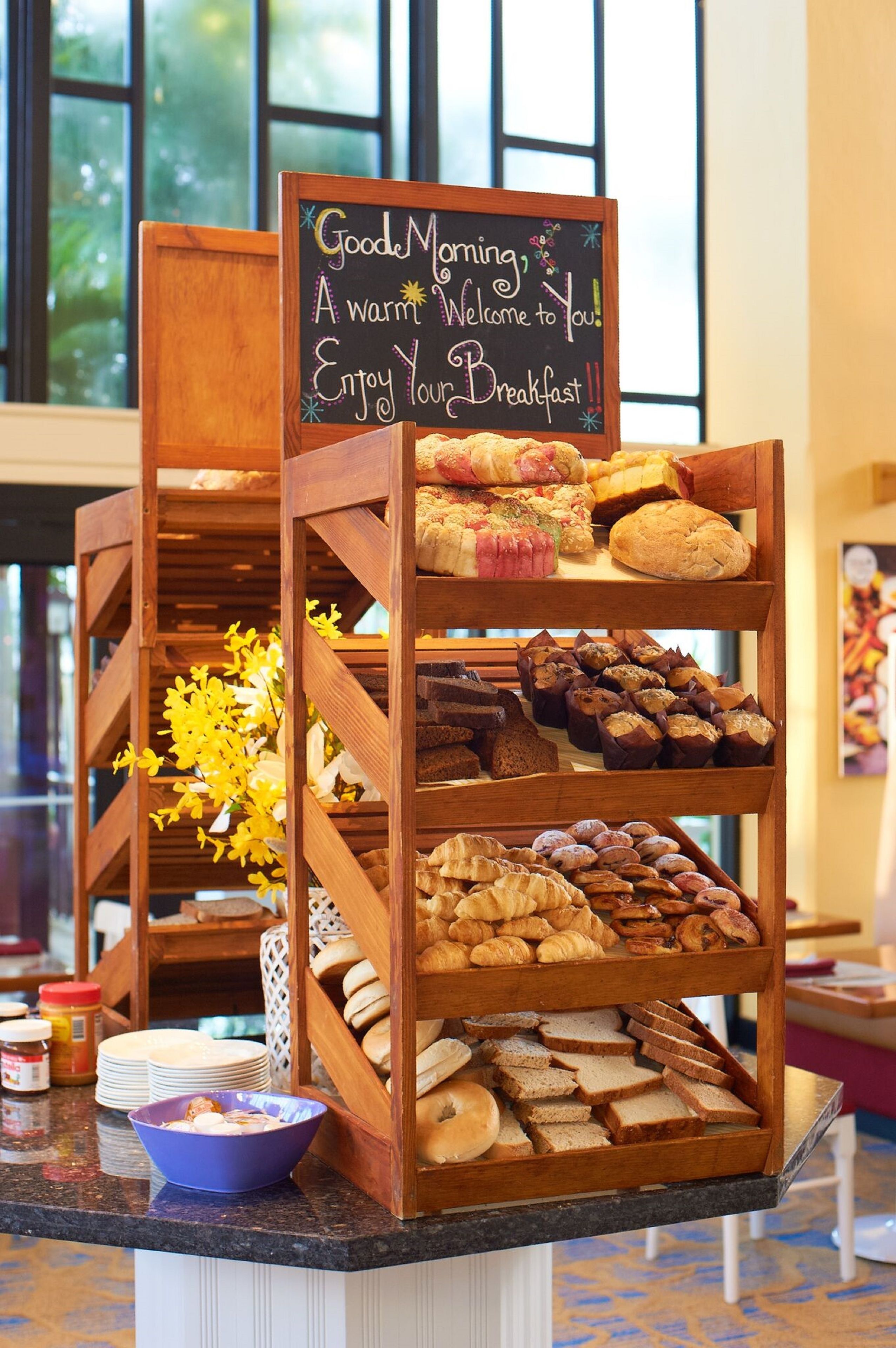A breakfast buffet with various pastries and bread, a sign welcoming guests, and a vase of yellow flowers.