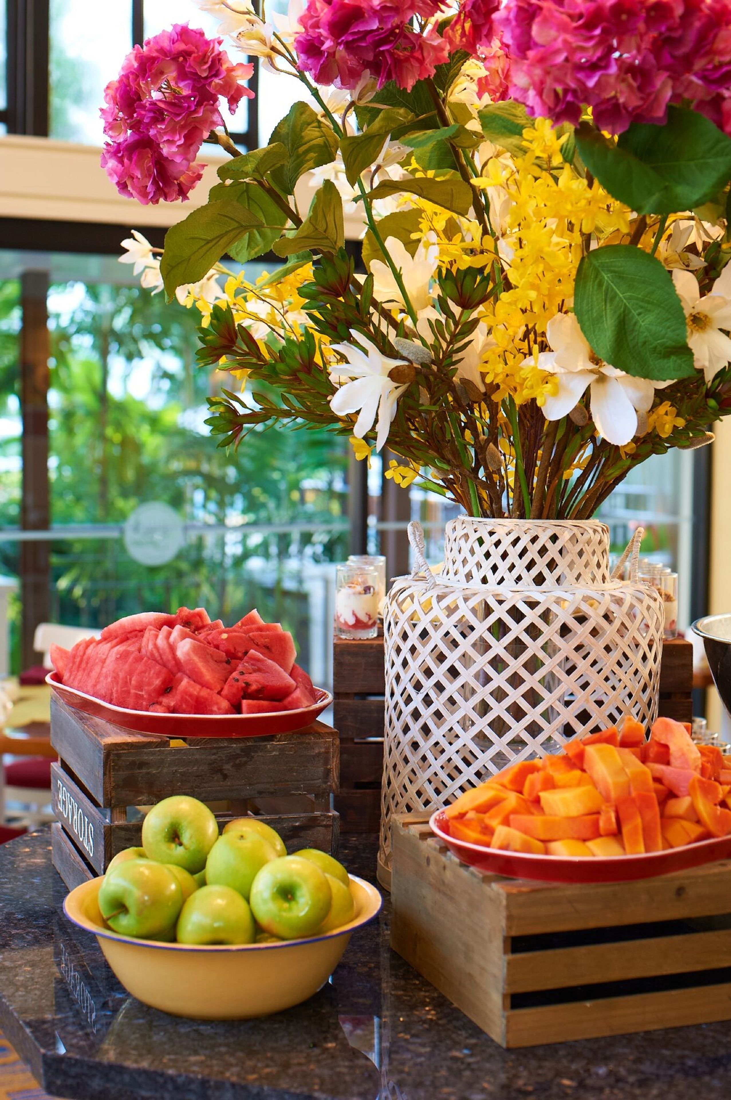 A fruit and flower arrangement on a counter with a window view.