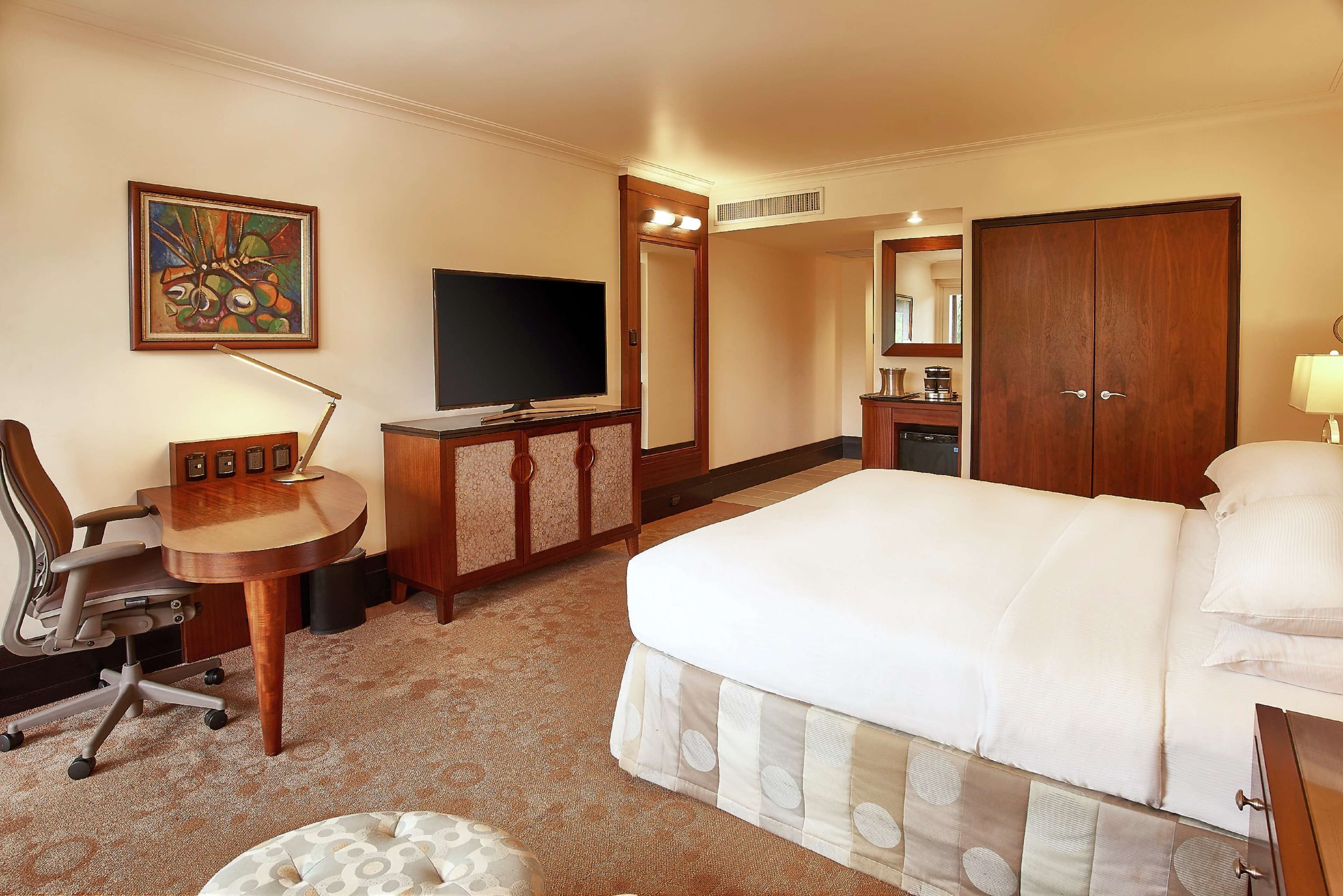A hotel room with a bed, a desk with a chair, a television, a wardrobe, and a bathroom mirror.