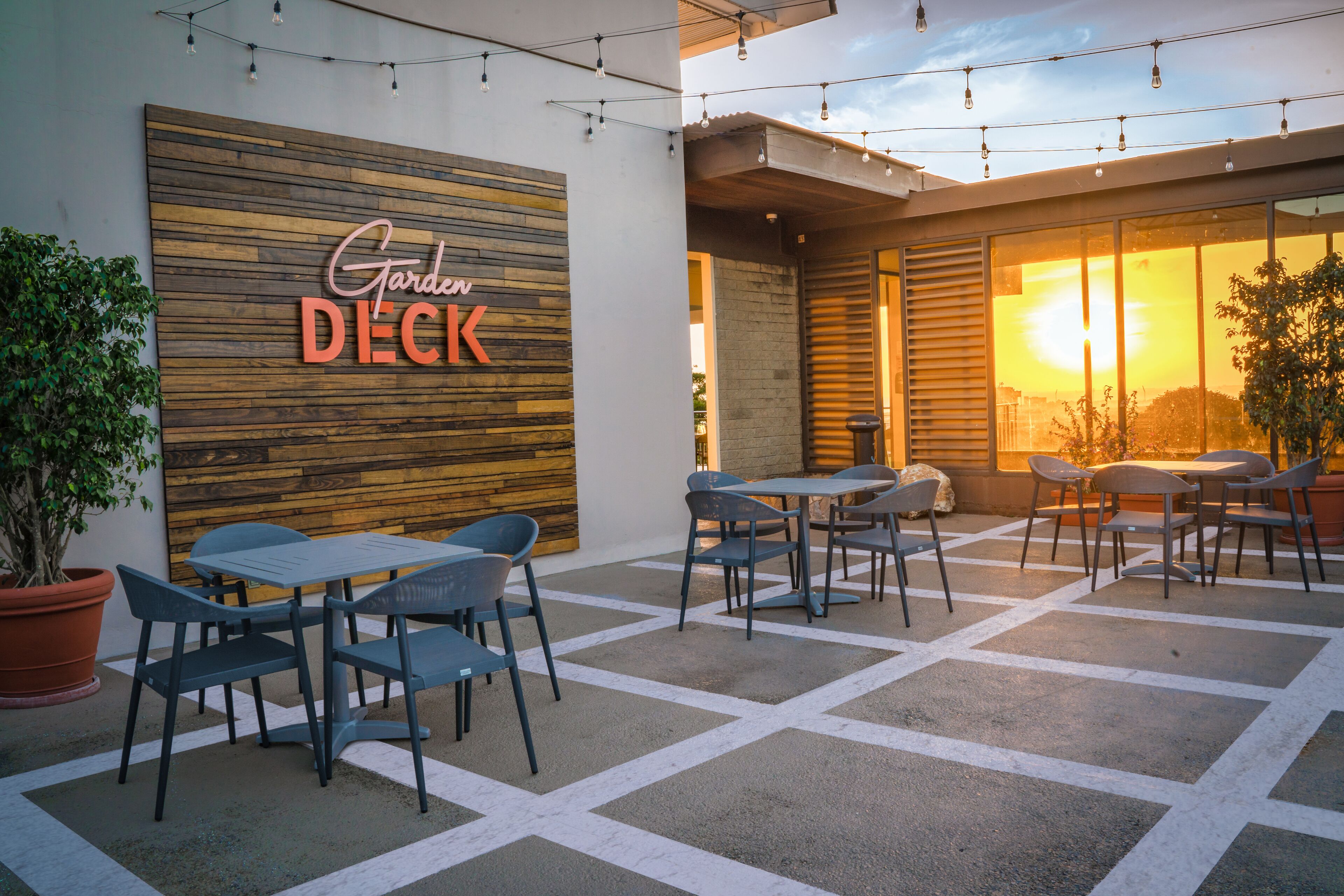 Un espace extérieur aménagé en terrasse, avec des tables et des chaises, un mur en bois sur lequel on peut lire l’inscription « Terrasse du jardin », et une vue sur le coucher du soleil à travers de grandes fenêtres.