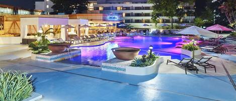 A nighttime pool area with lounge chairs and umbrellas, surrounded by buildings and greenery.