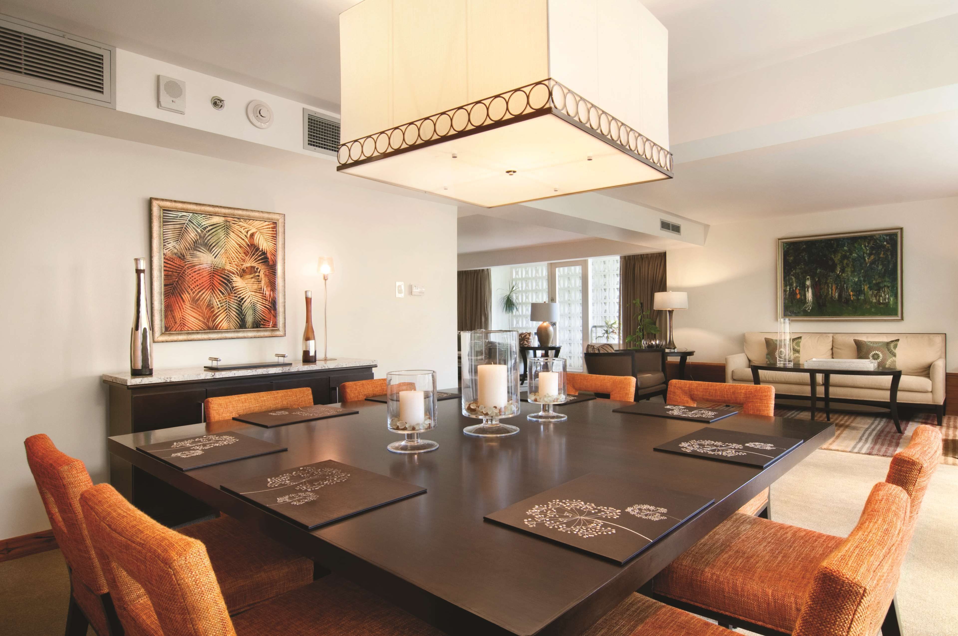 A spacious dining area with a large rectangular table, orange upholstered chairs, and a central chandelier.