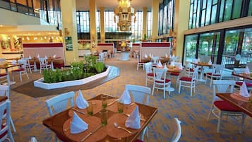 A spacious hotel lobby with multiple dining areas, large windows, and a central planter with greenery.