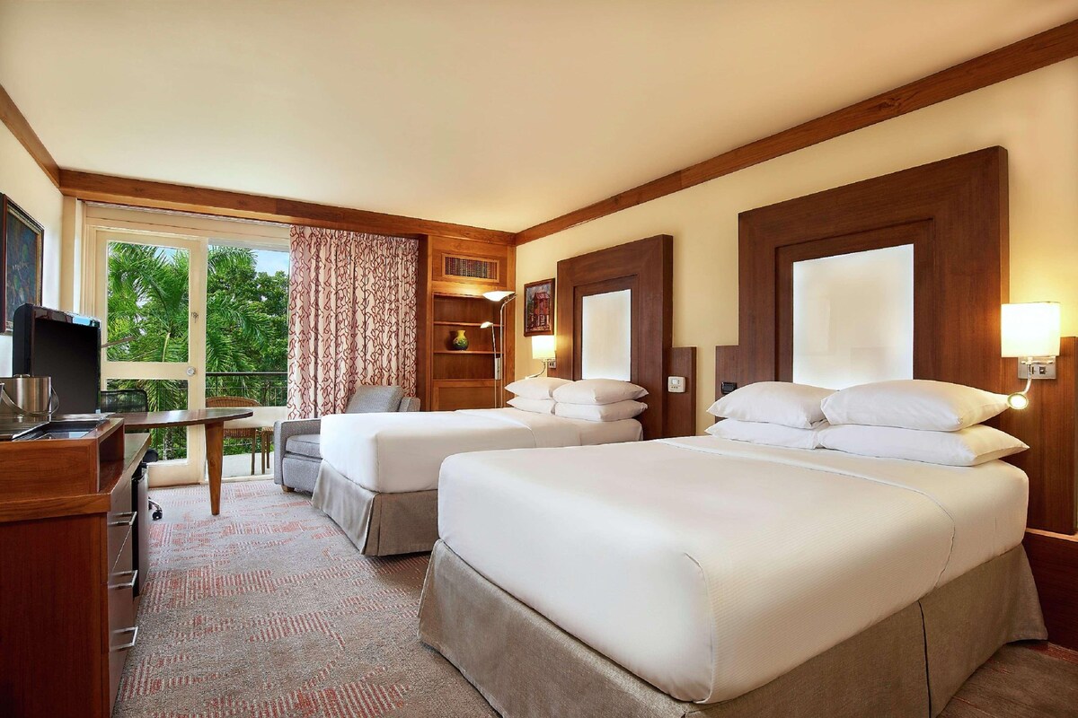 A hotel room with two beds, a desk, a chair, a TV, and a balcony with a view of greenery.