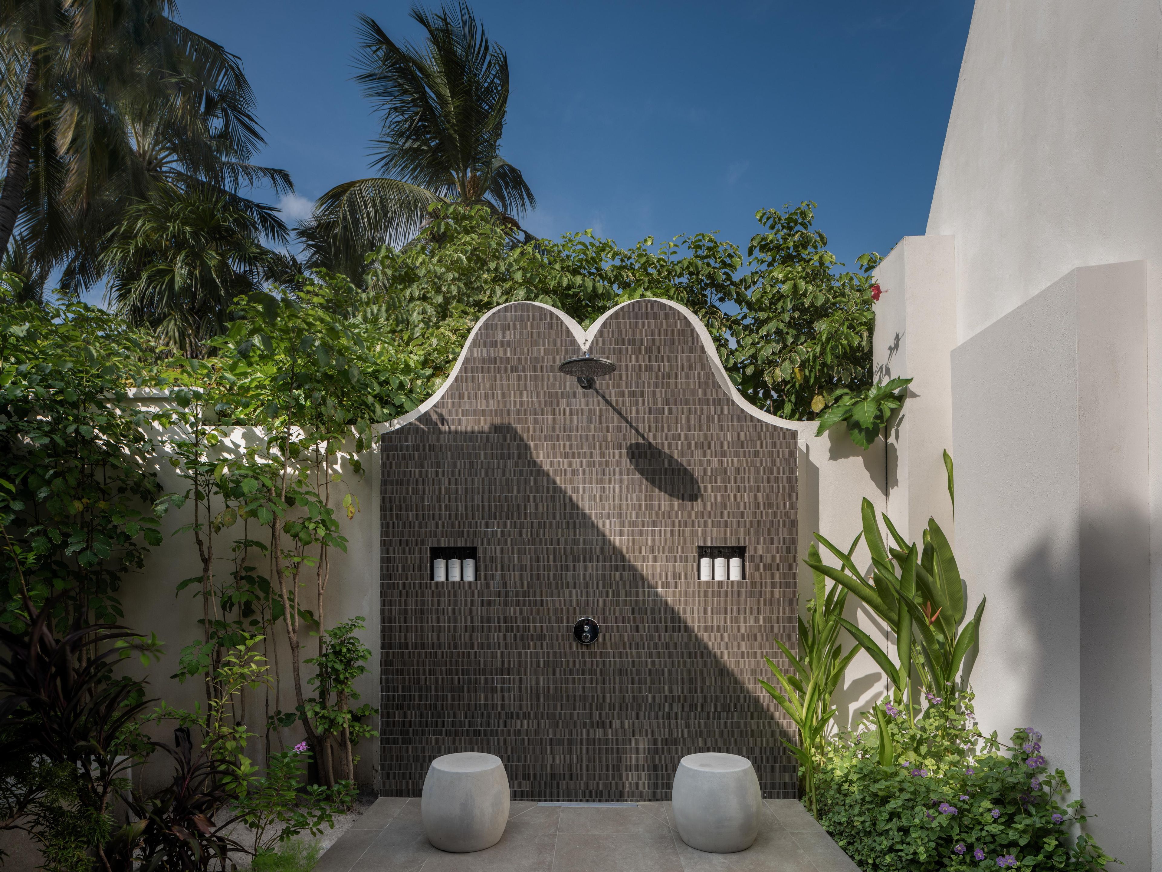 A modern outdoor shower area with a tiled wall, two large stone seats, and lush greenery.