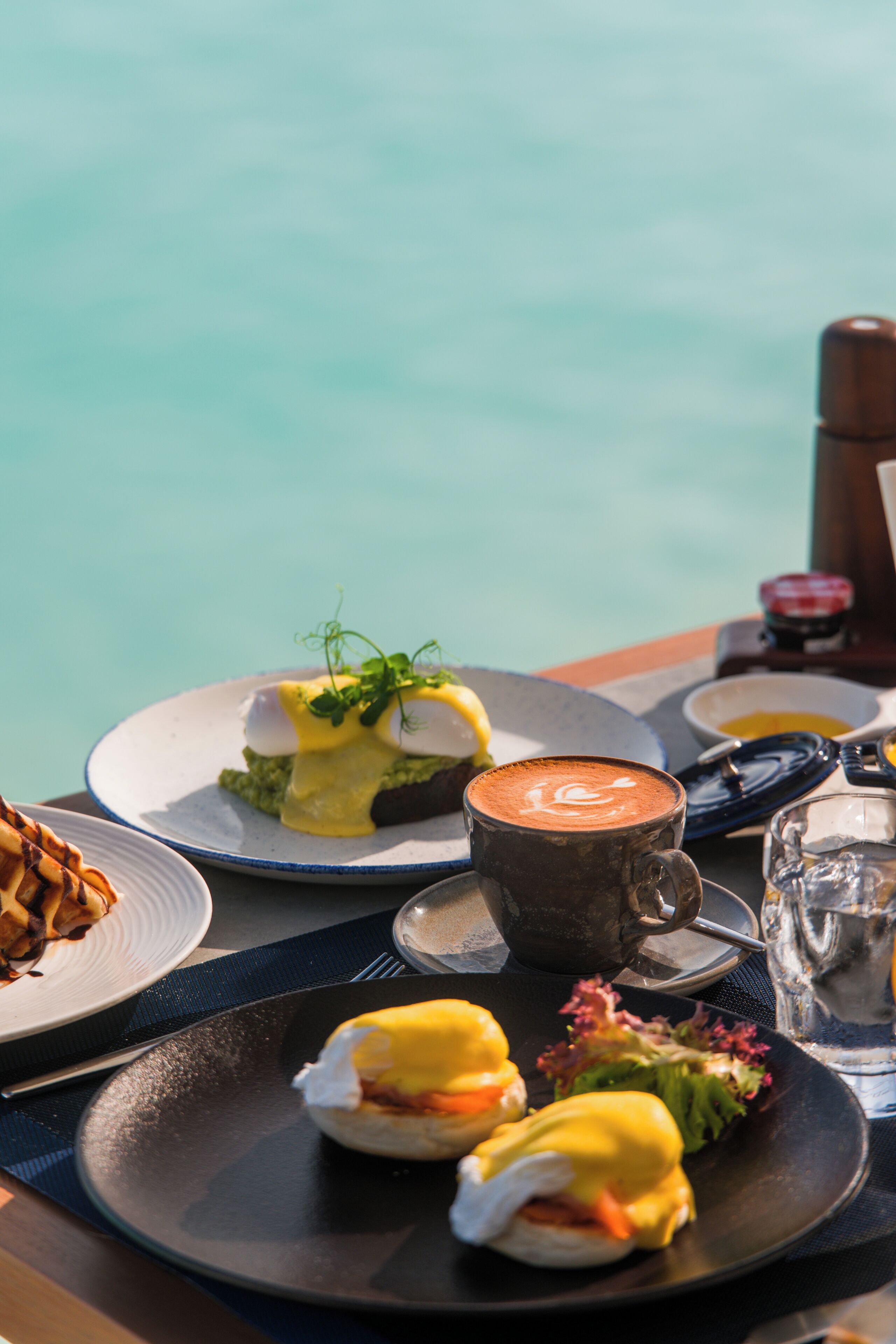 A table with breakfast items including eggs benedict, a cup of coffee, and a plate of food, overlooking a body of water.
