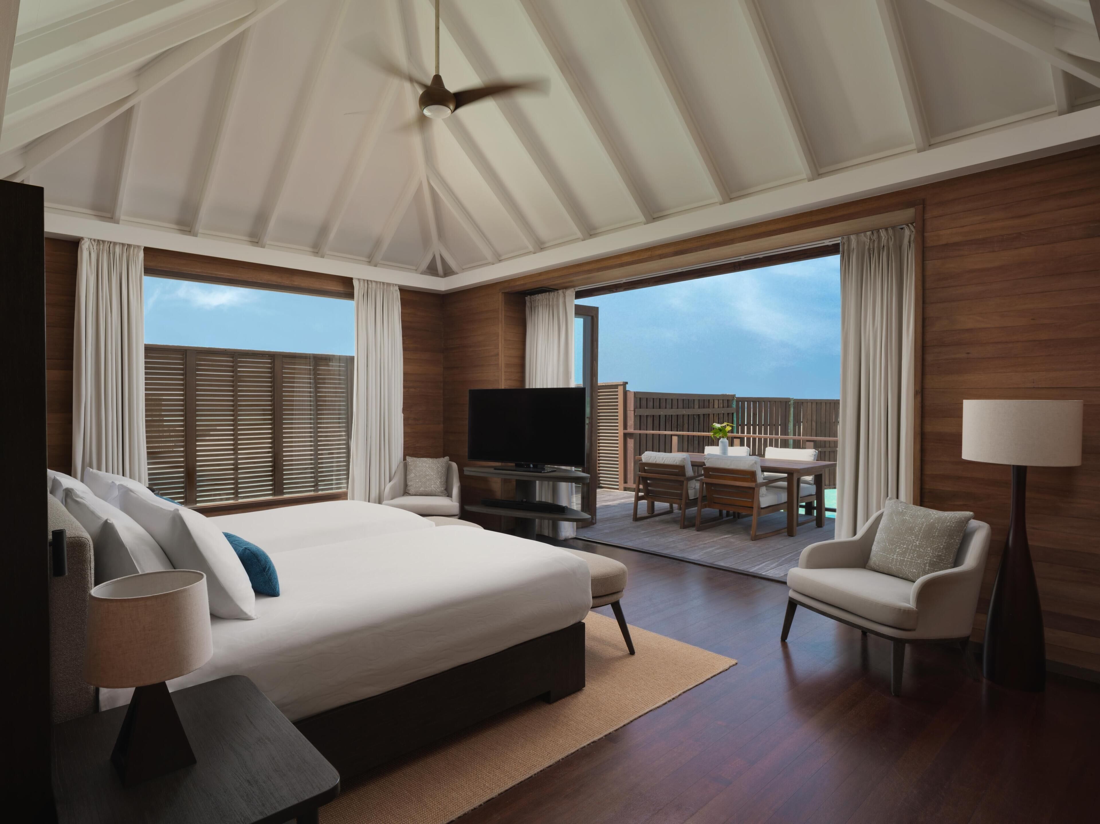A hotel room with a large bed, a TV, a balcony with a table and chairs, and a ceiling fan.
