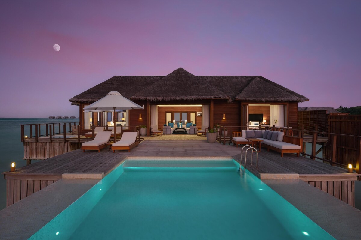 A resort with a pool, thatched-roof overwater bungalows, and a view of the ocean.