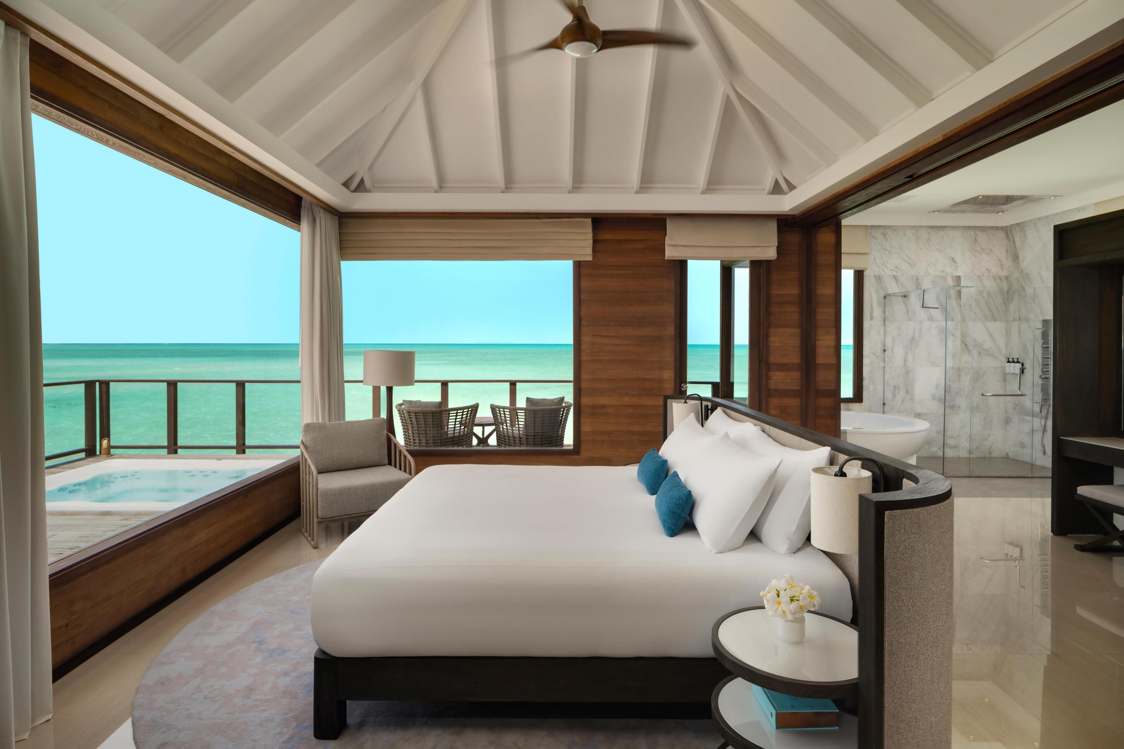 A modern hotel room with a large bed, a balcony with a view of the ocean, a bathroom with marble, and a ceiling fan.