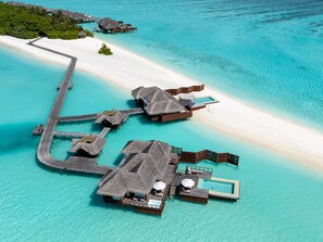An aerial view of overwater bungalows connected by walkways, with clear turquoise waters and a sandy beach.