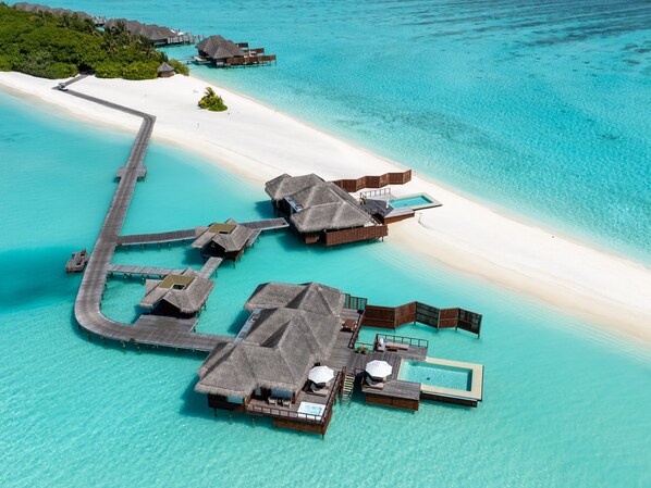 An aerial view of overwater bungalows connected by walkways, with clear turquoise waters and a sandy beach.
