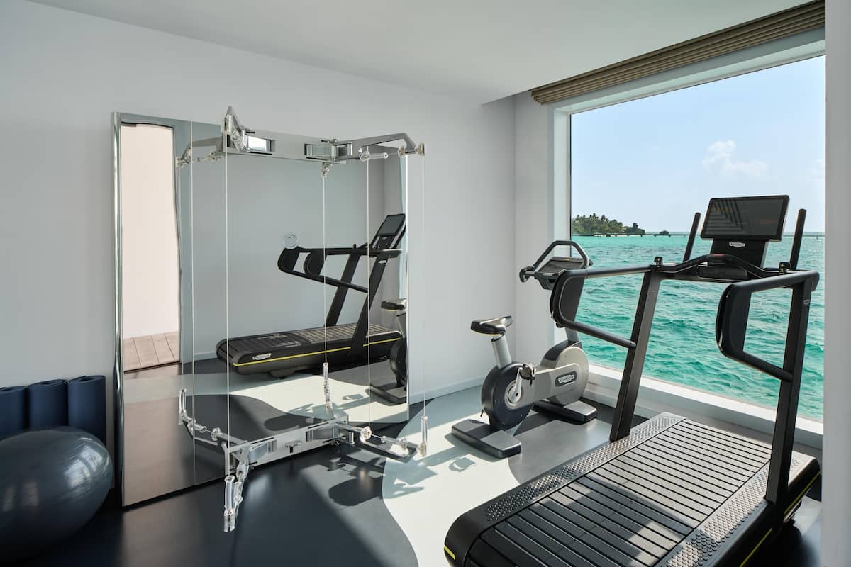 Uma academia moderna com esteira, elíptica e bola de exercícios, com vista para o mar azul-turquesa.