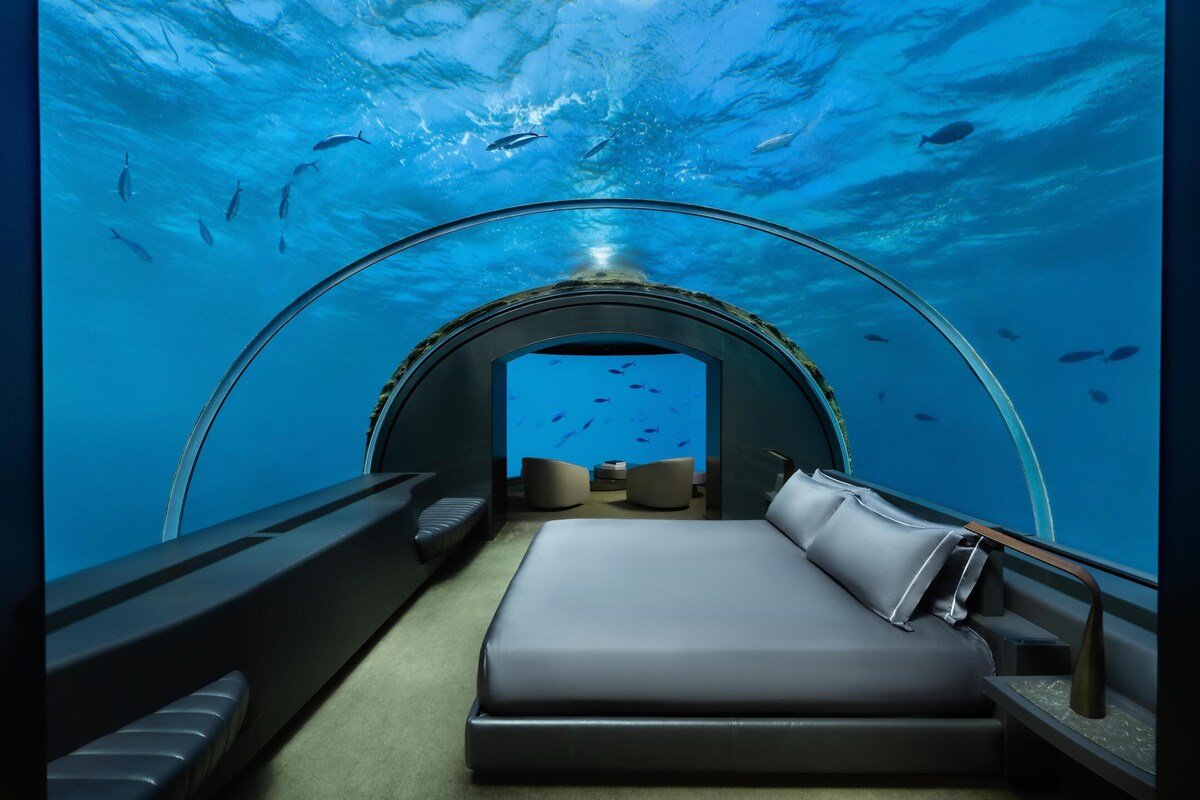 An underwater room with a bed, seating area, and a view of fish swimming outside.