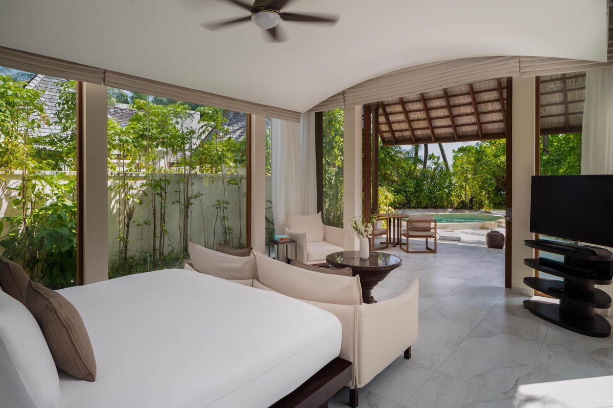 A spacious room with a bed, a seating area with a table, a TV, and large windows offering a view of greenery.