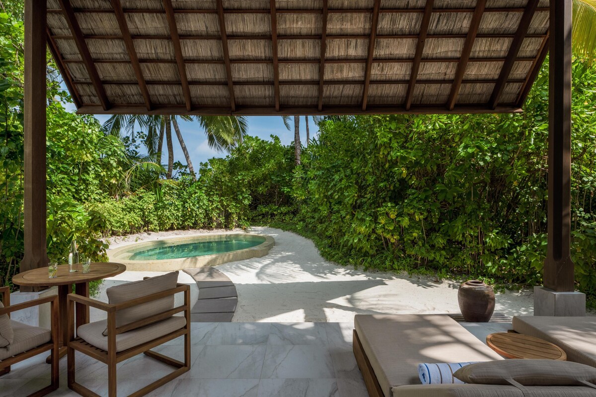 A covered outdoor seating area with a pool, surrounded by lush greenery.
