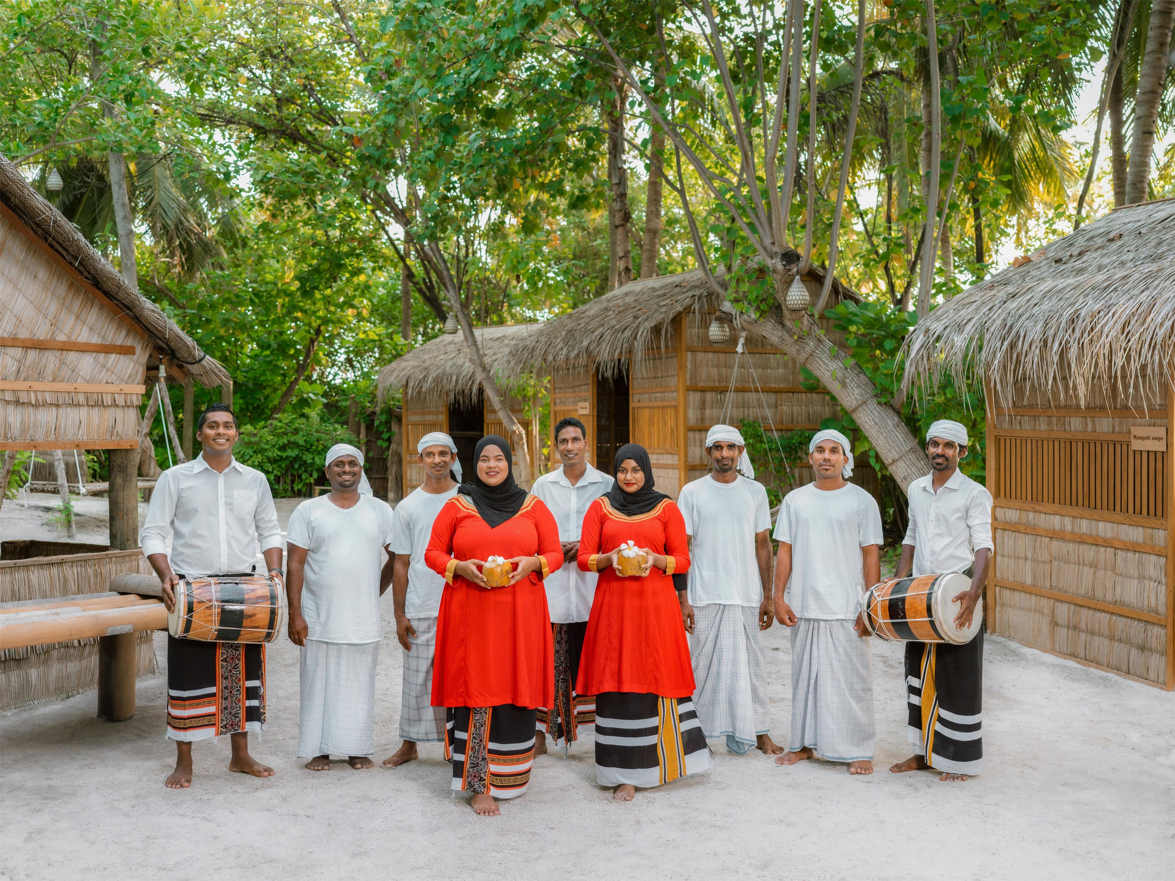 A group of people standing in a tropical setting with thatched-roof huts and lush greenery in the background.