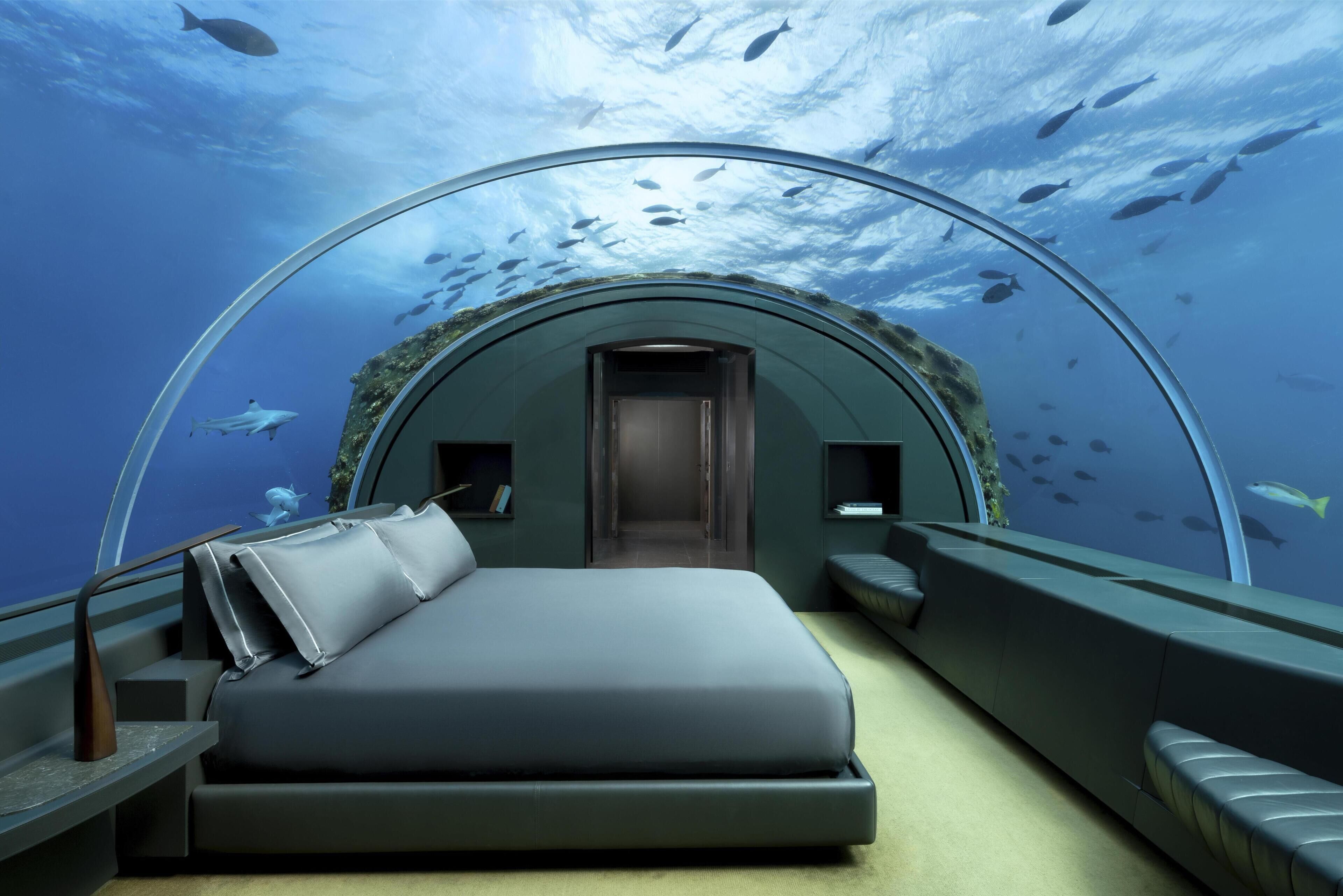 A transparent dome room with a bed, seating area, and a view of fish swimming outside.
