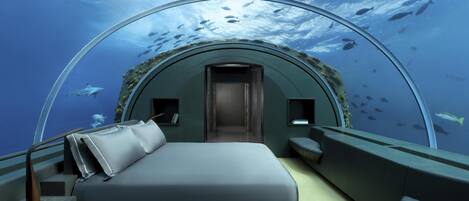 A transparent dome room with a bed, seating area, and a view of fish swimming outside.