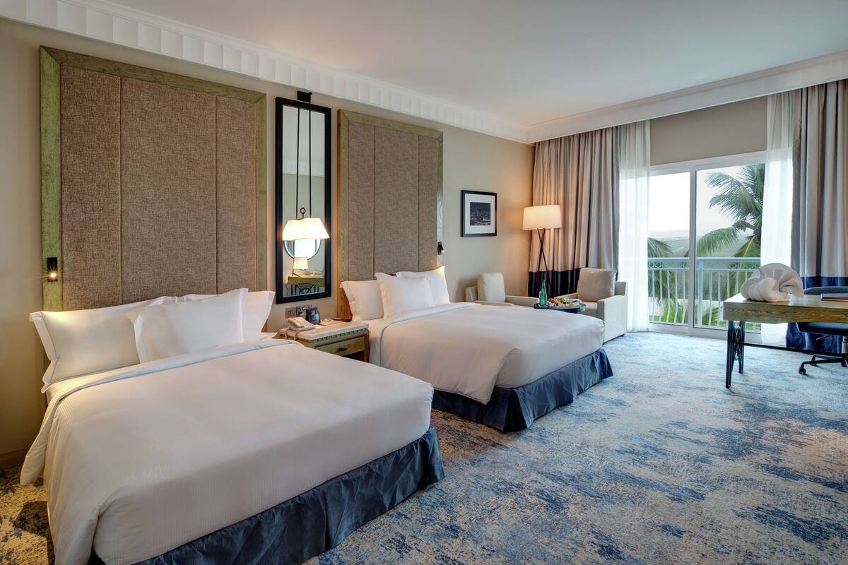 Habitación de hotel con dos camas, un escritorio, una silla, un ventanal con vistas y un balcón.