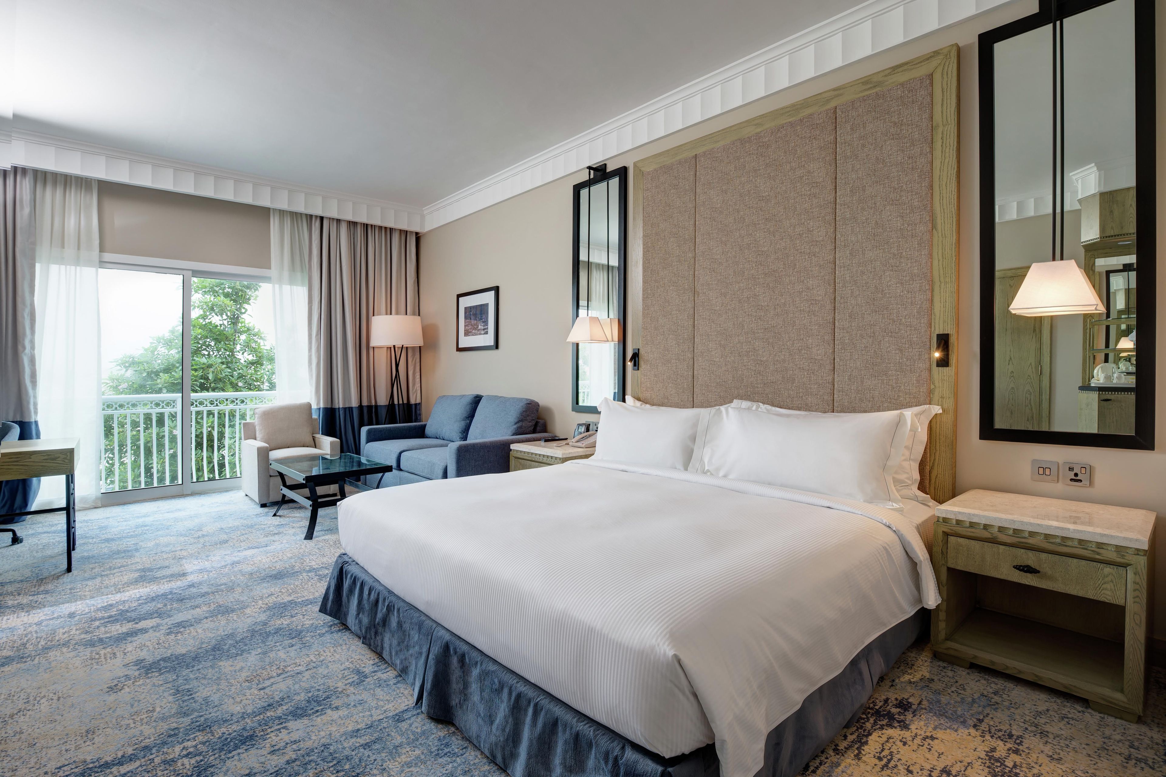 Quarto de hotel com uma cama grande, escrivaninha, cadeira, sofá, varanda com cortinas e um espelho amplo.