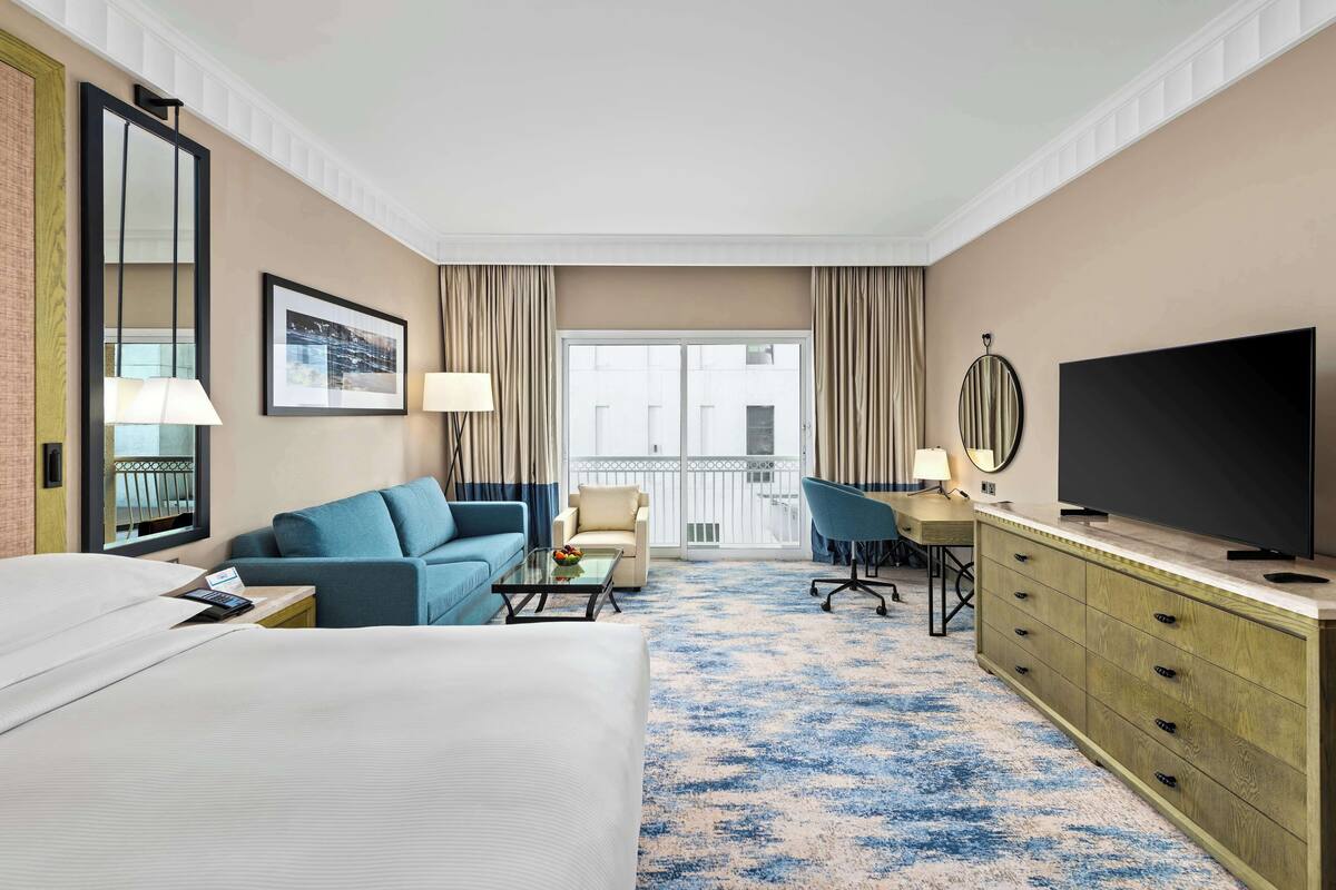 Ett hotellrum med en säng, en blå soffa, ett skrivbord med en tv, en stol och ett stort fönster med gardiner.