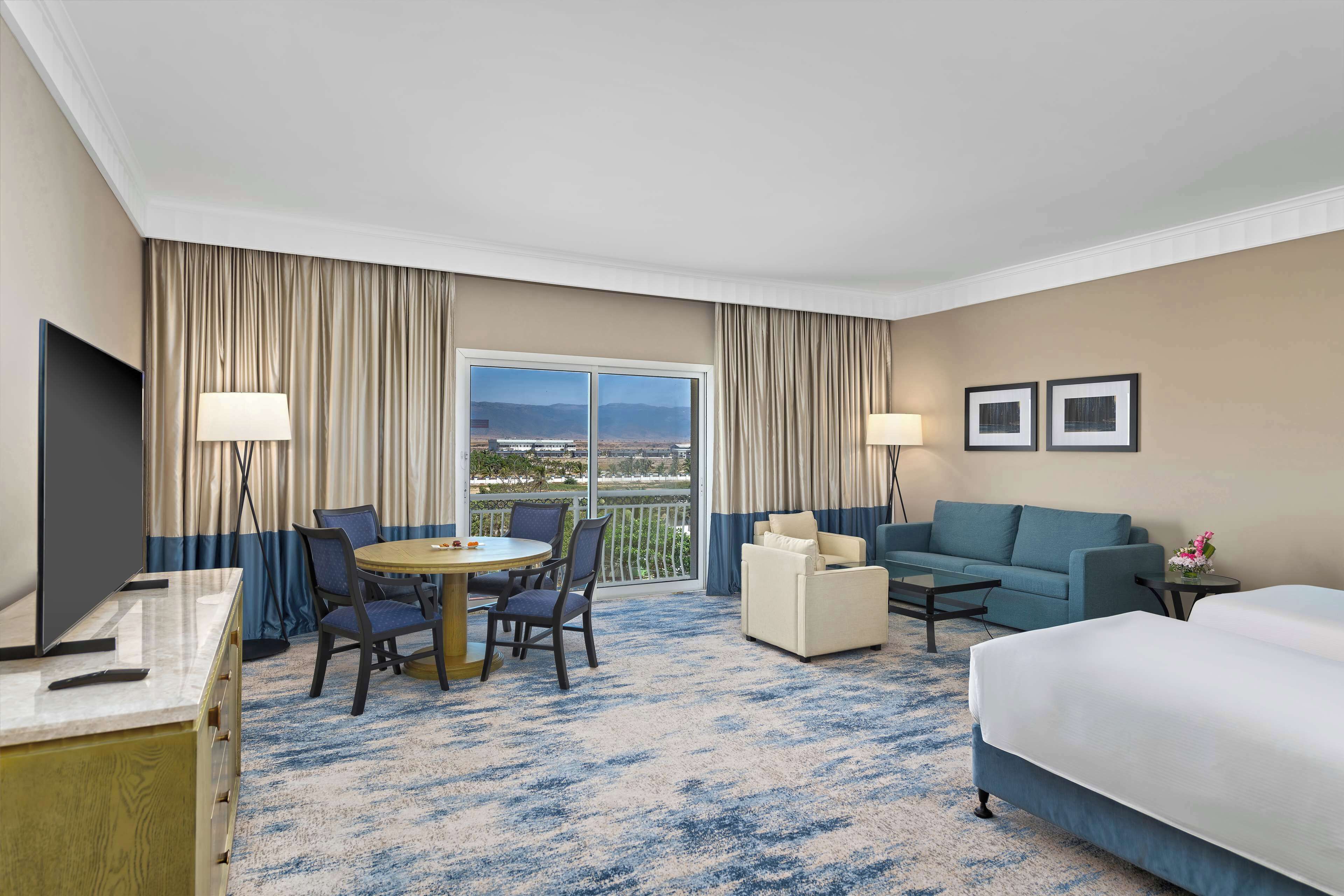 A hotel room with a bed, a desk, a chair, a table, and a balcony with curtains.