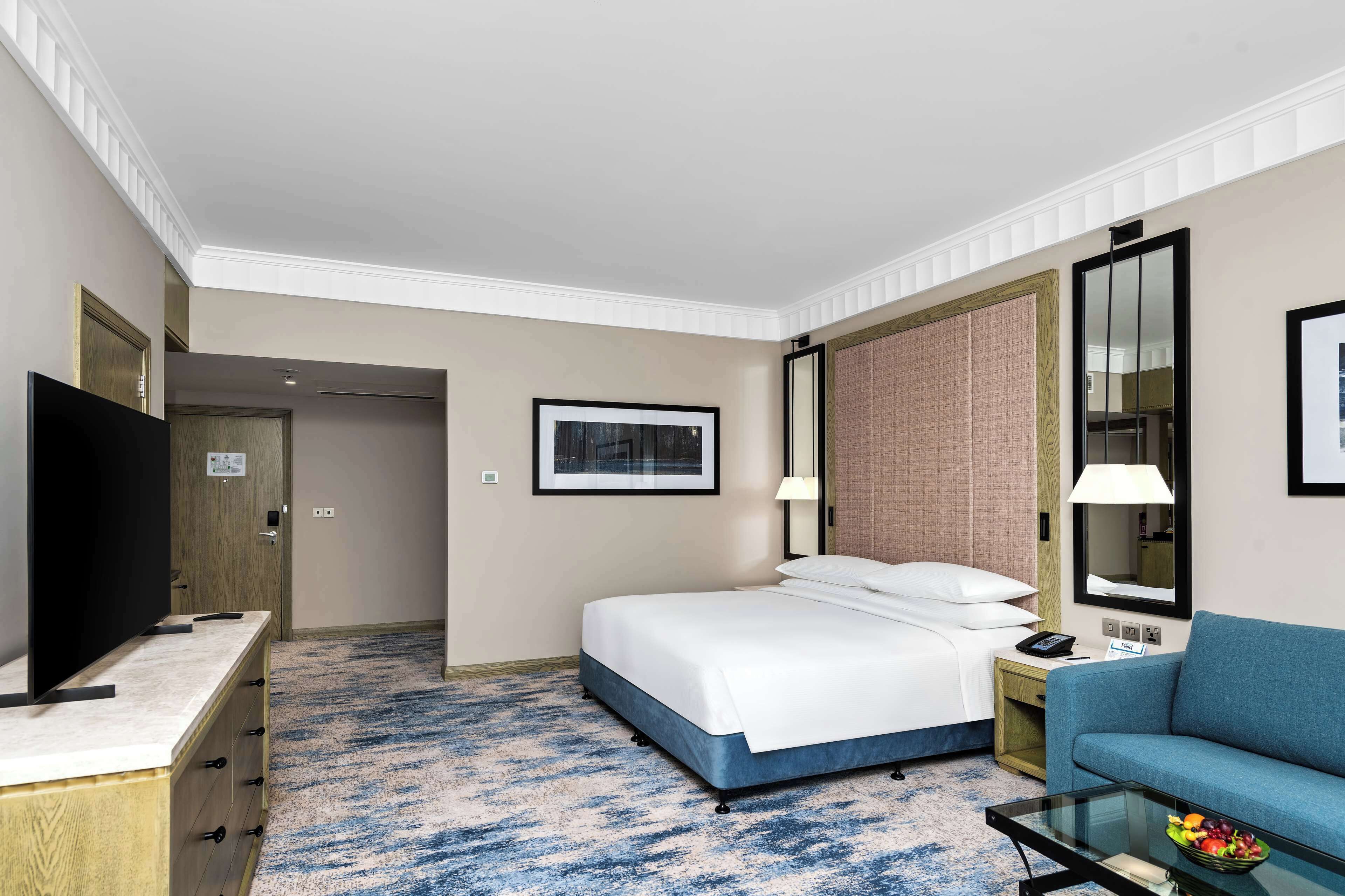 Habitación de hotel con cama, televisión, sofá, una mesita de vidrio con fruta y un espejo grande.