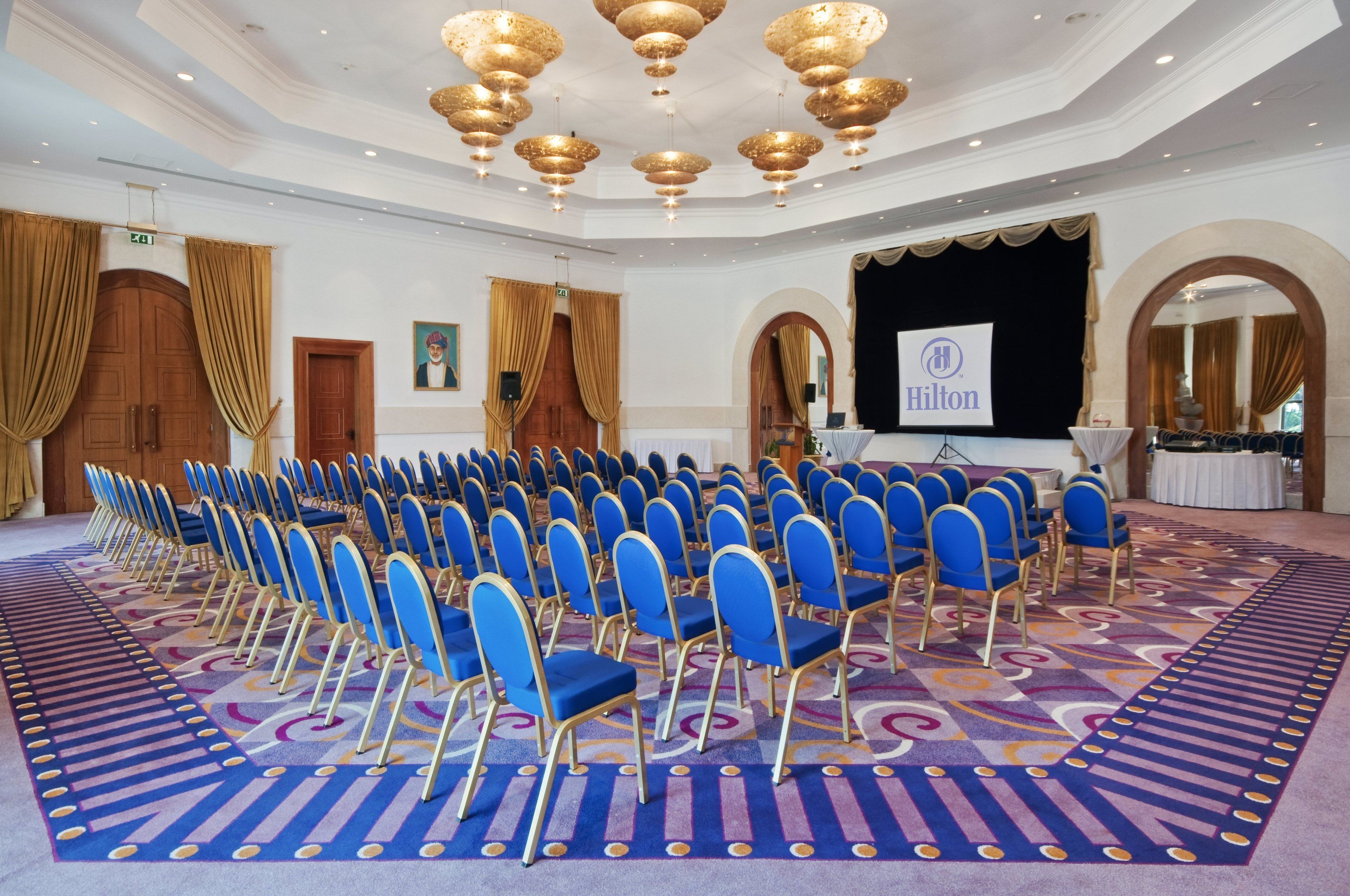 A large conference room with rows of blue chairs, a stage with a 'Hilton' banner, and ornate ceiling chandeliers.