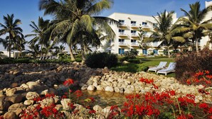 Un complexe hôtelier doté d’une piscine entourée de rochers et de fleurs rouges, de palmiers et d’un bâtiment agrémenté de balcons.
