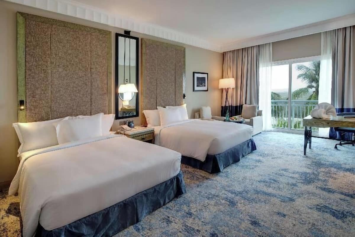 Une chambre d’hôtel avec deux lits, un grand miroir, un bureau avec une lampe et une vue sur des palmiers.
