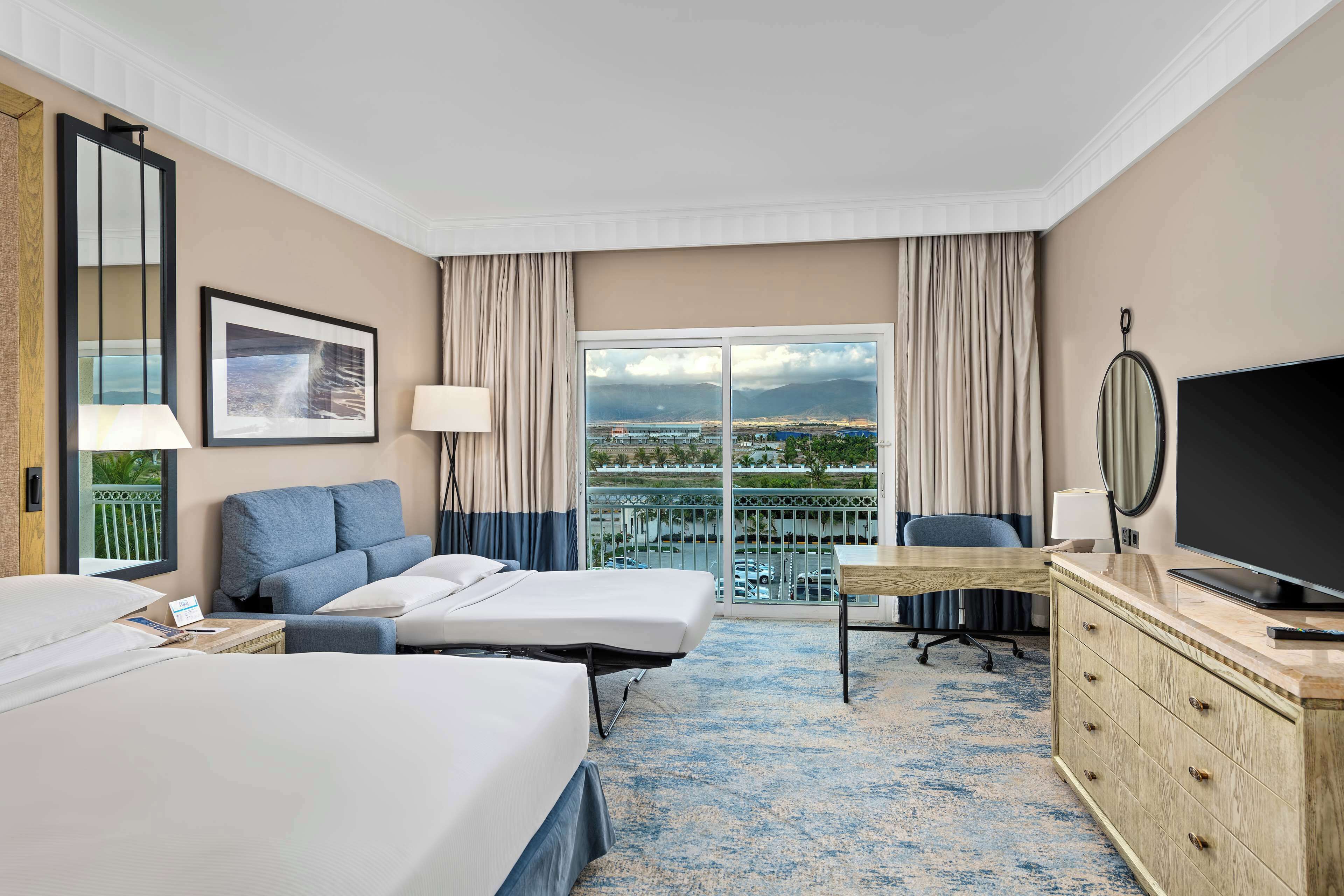 Ein Hotelzimmer mit zwei Betten, einem Schreibtisch, einem Fernseher und einem Balkon mit Blick auf die Stadt.