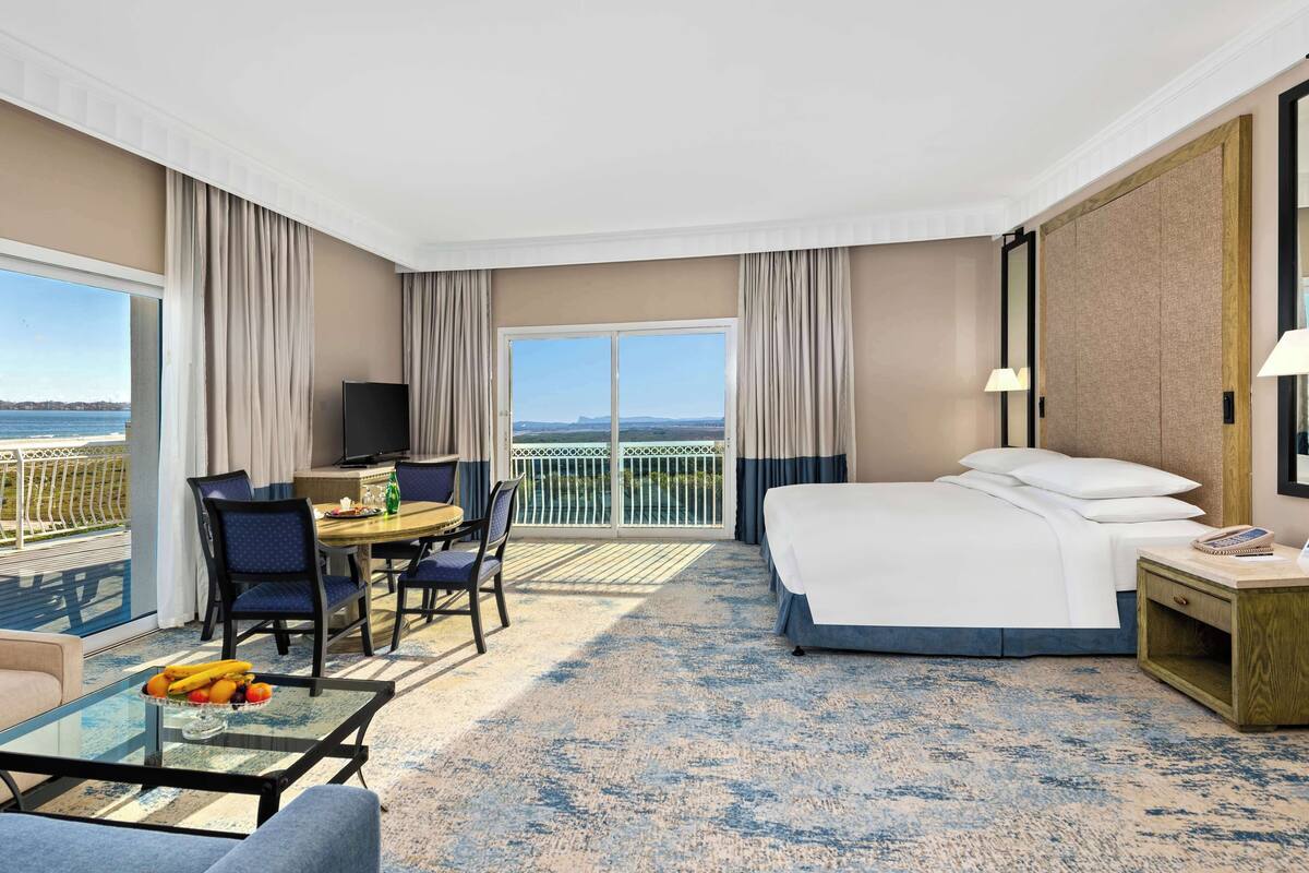 Habitación de hotel con balcón, una cama, un escritorio con silla, una mesa de vidrio con fruta y vista al mar.