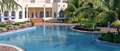 A large pool with a poolside area, palm trees, and a building with arched windows and a tower.