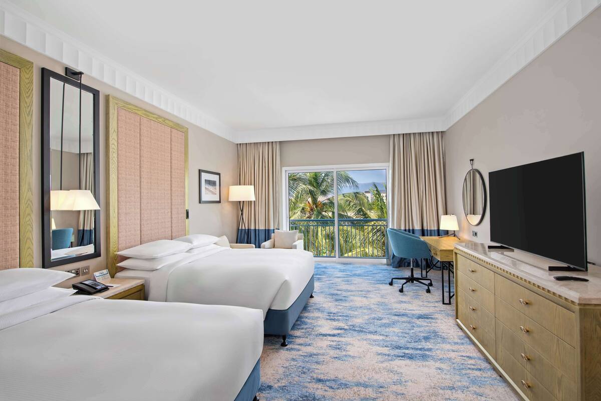 Habitación de hotel con dos camas, un escritorio, televisión y balcón con vista a palmeras.