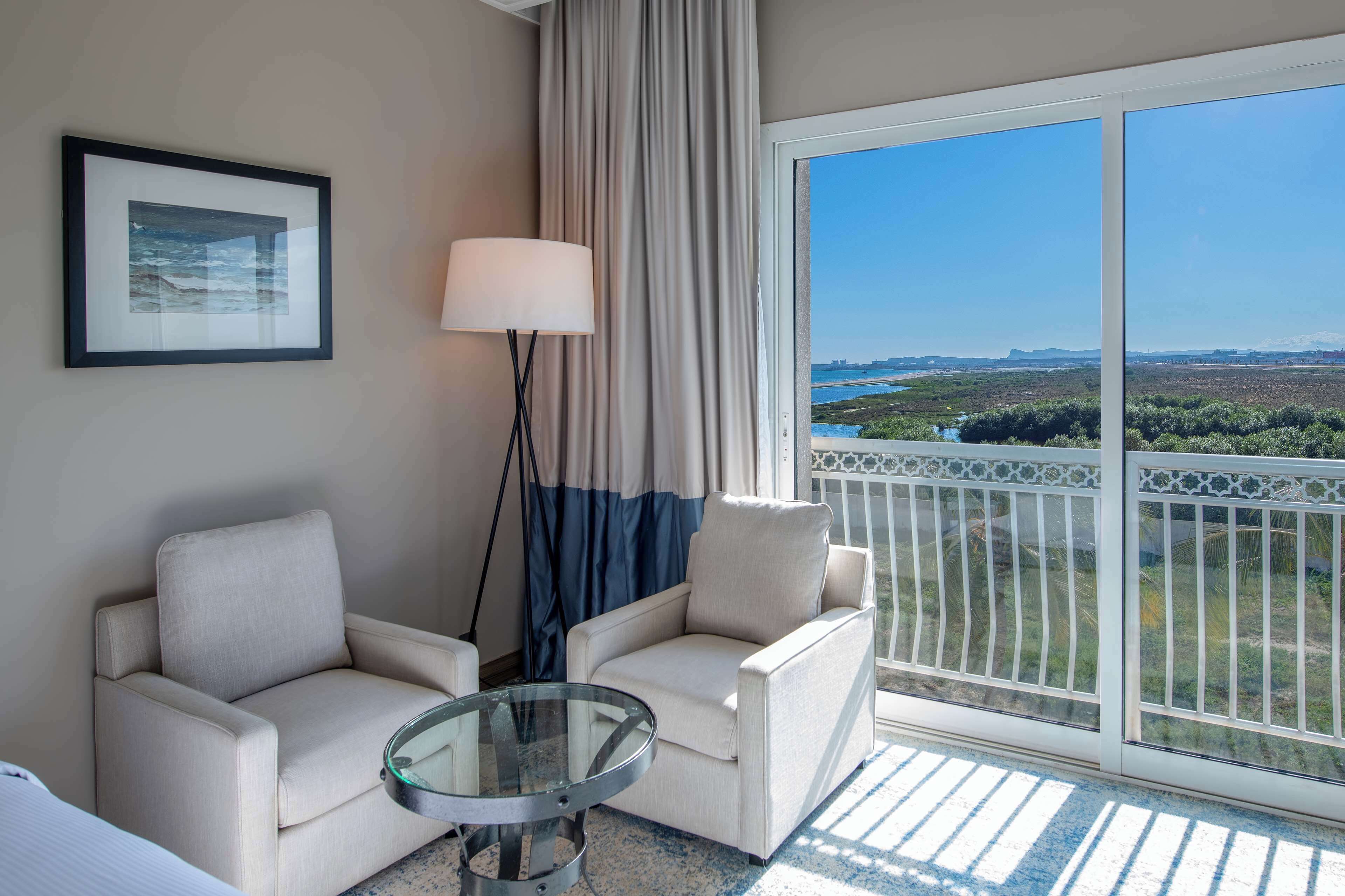 A hotel room with a balcony, a framed picture on the wall, a lamp, and two armchairs.