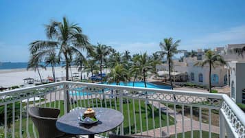 A balcony with wicker chairs and a table, overlooking a pool and beach area with palm trees and a clear sky.