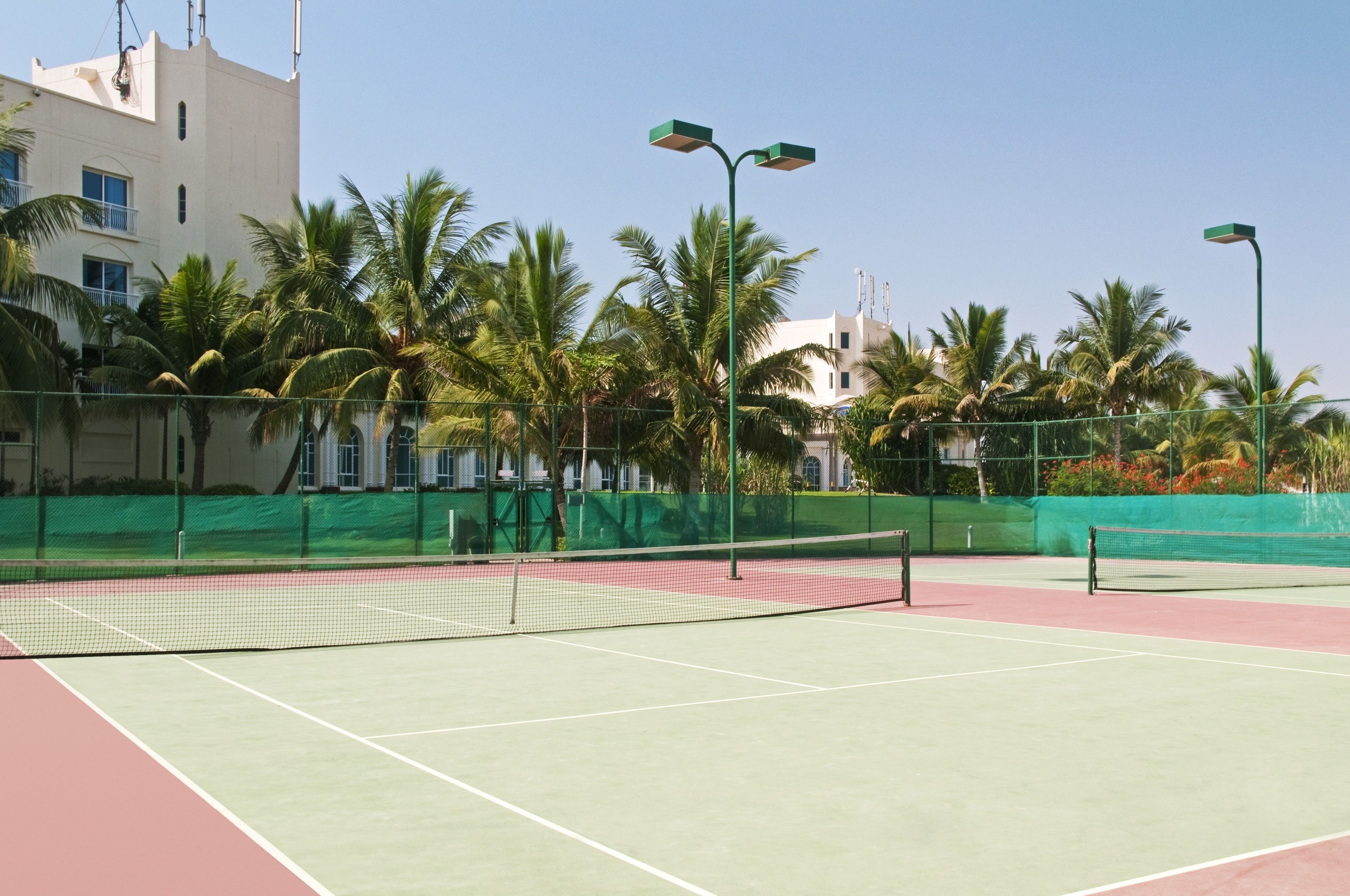 An outdoor tennis court with a net, surrounded by palm trees and a building in the background.