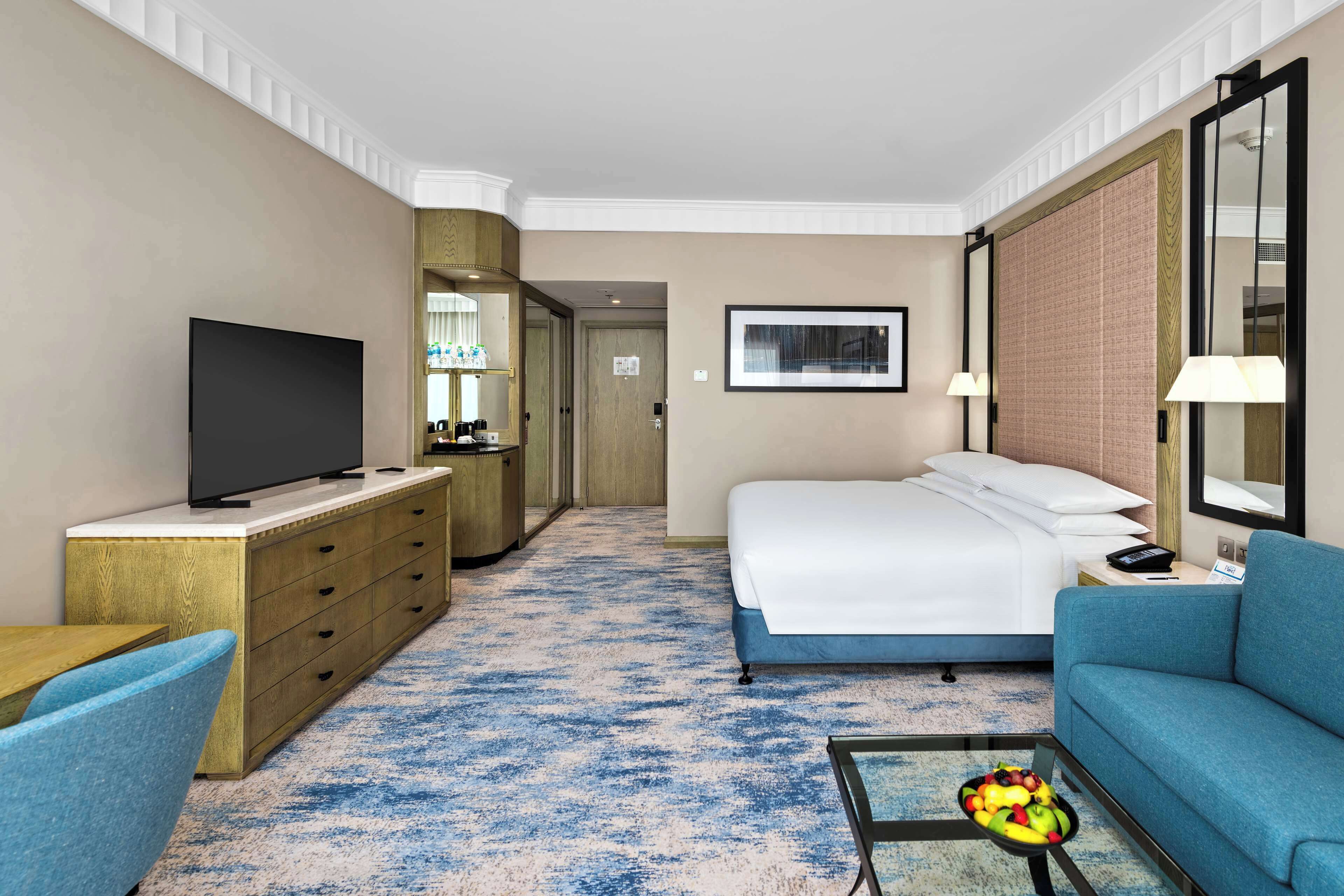 Una habitación de hotel con una cama, televisión, escritorio con computadora, sofá azul y mesa de centro con fruta.