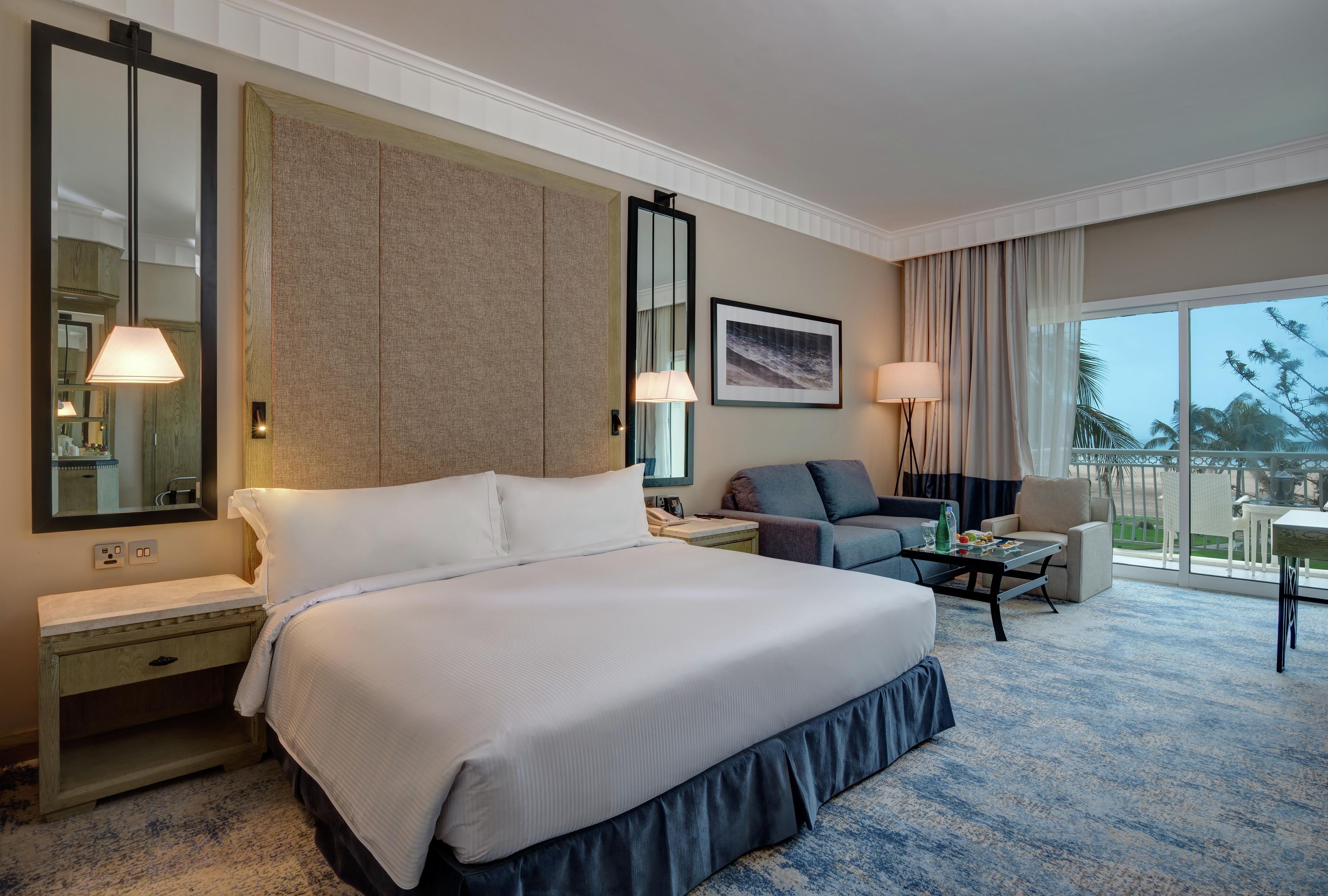 A hotel room with a large bed, a sitting area with a sofa and armchairs, a dining table, and a balcony with a view.